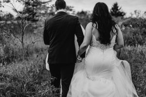 Black and white photo of a couple walking through a field on their wedding day, viewed from behind.