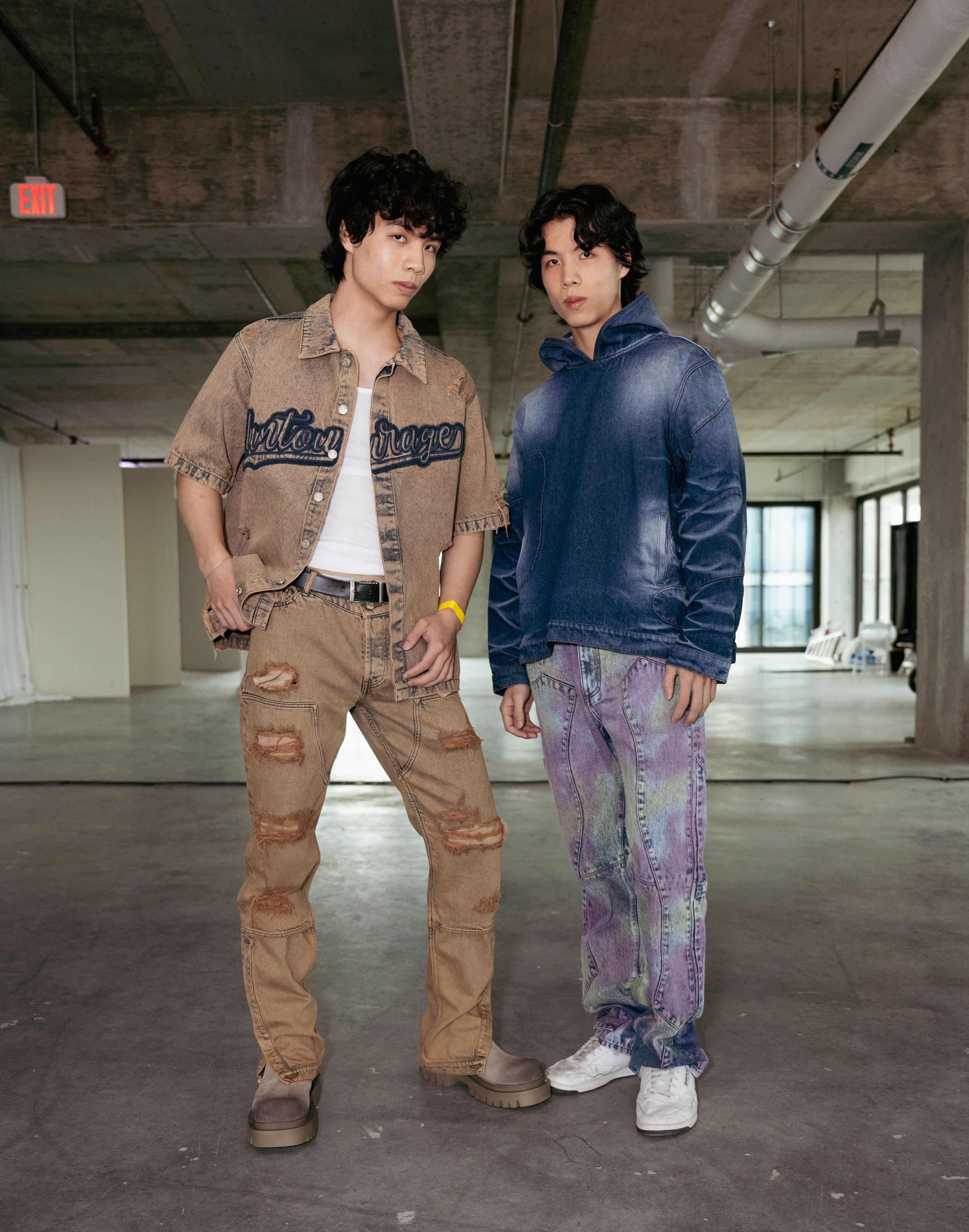 Two models in casual streetwear with denim and tan utility clothing in an empty industrial space.