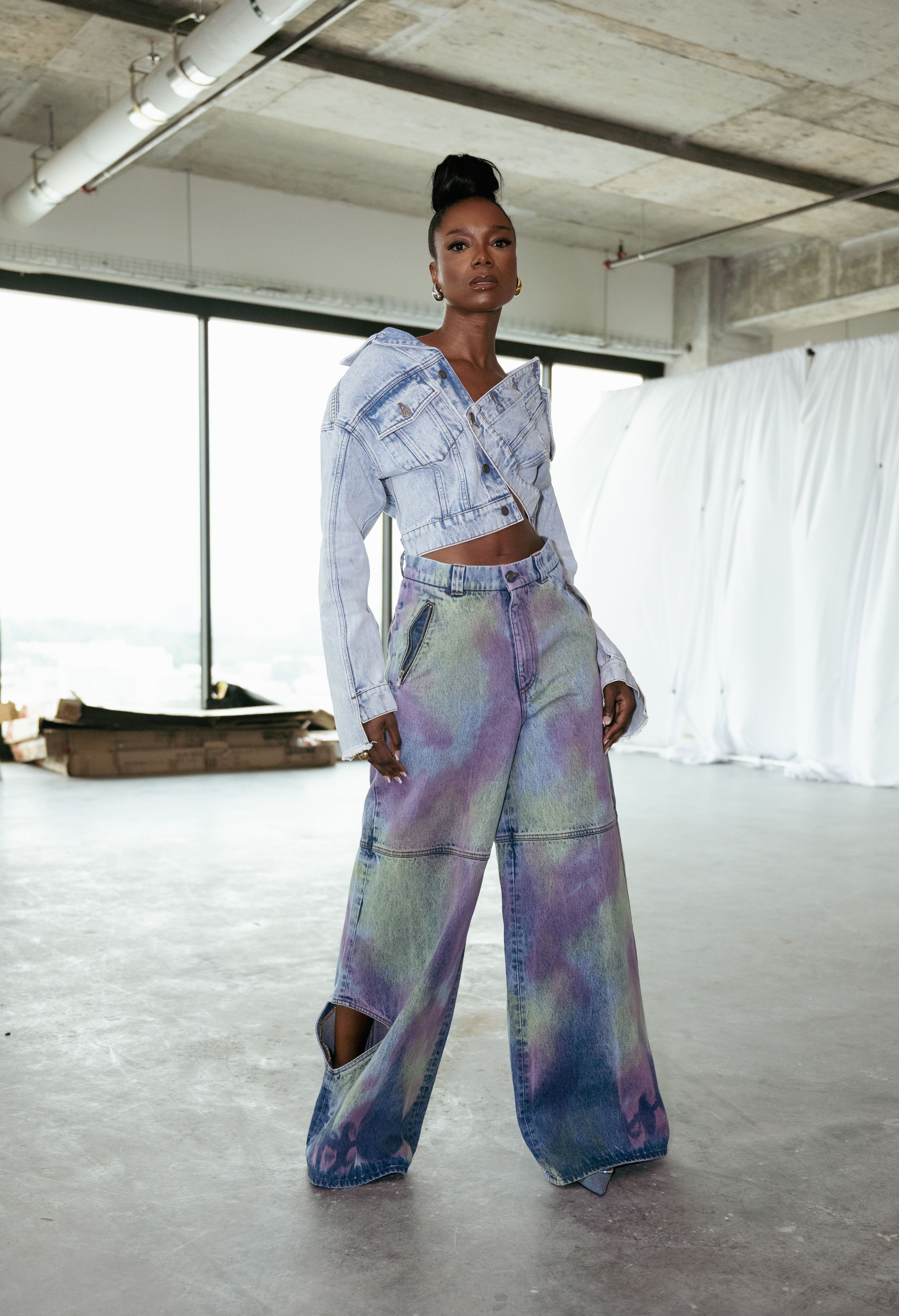 Fashion model in tie-dye wide leg jeans and cropped denim jacket poses in industrial space.