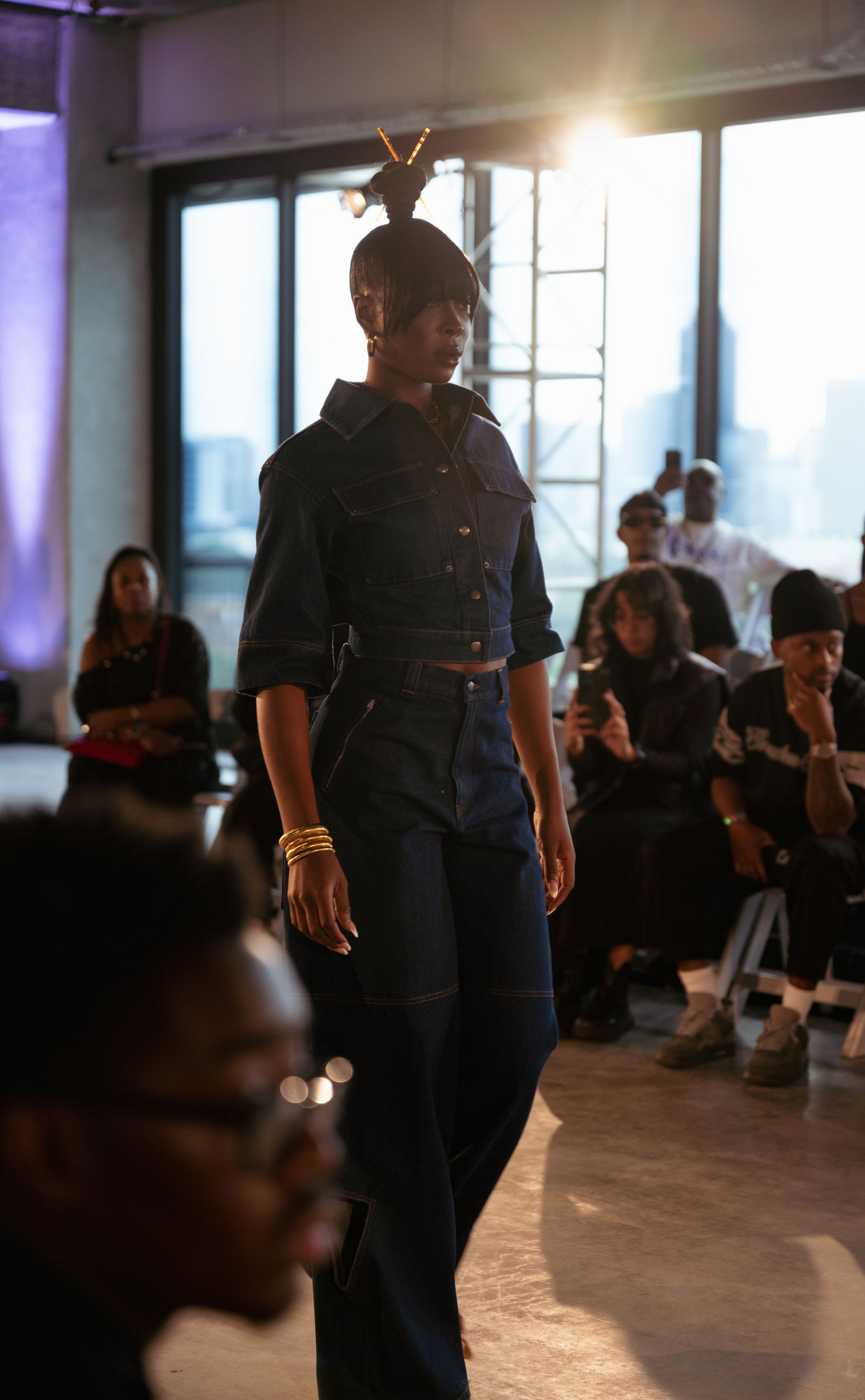 Fashion model walks runway in all-denim ensemble with high ponytail under dramatic lighting.