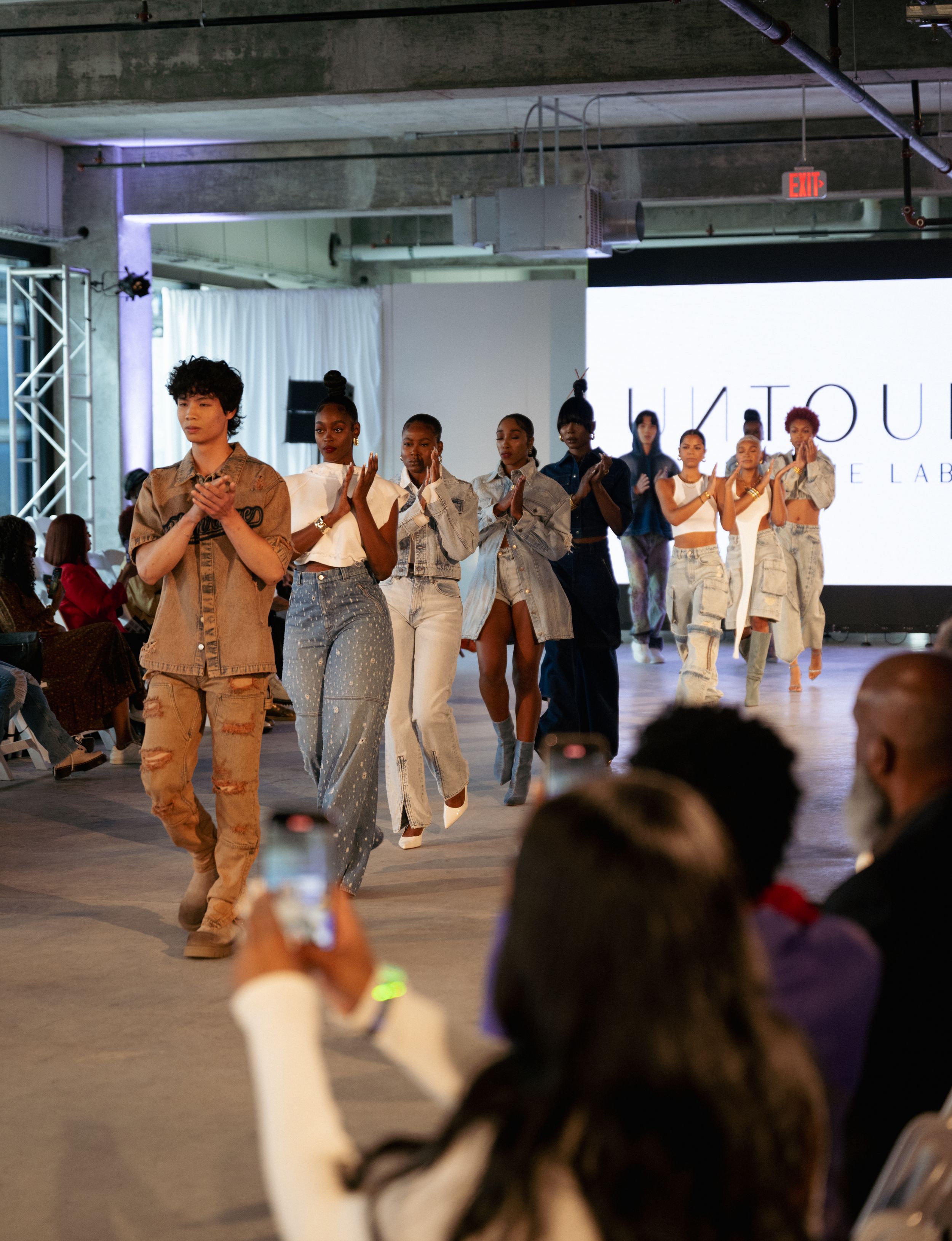 Models walk runway finale in neutral toned casual wear collection at indoor fashion show.