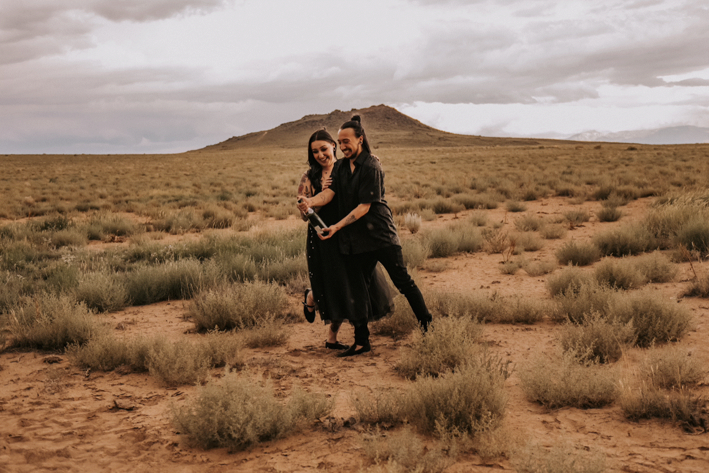 A couple embraces in a desert landscape with sage brush and mountains in the distance. LGBT engagement photographer Albuquerque. ABQ photos.
