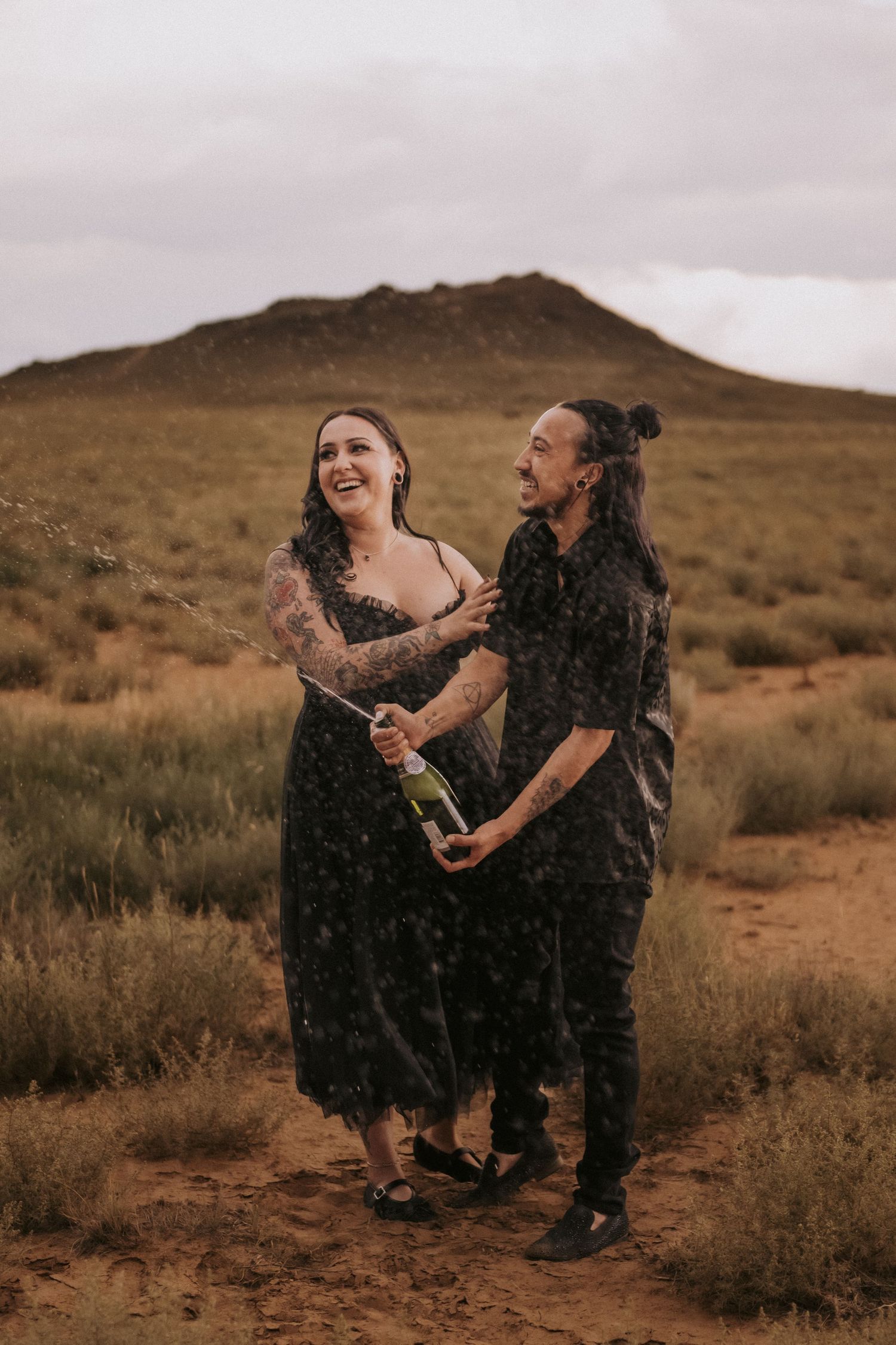 A couple laughs and pops champagne in the desert. Engagement photo locations in New Mexico. Affordable albuquerque couples photographer.