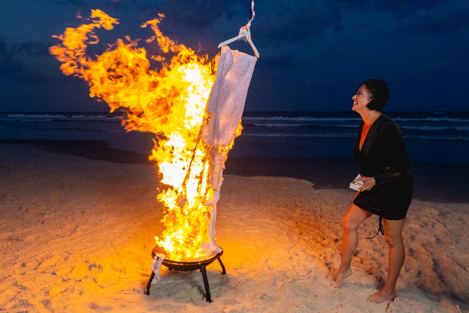 A person stands on a beach at night watching a large fire burn with dramatic orange flames lighting up the dark scenery.