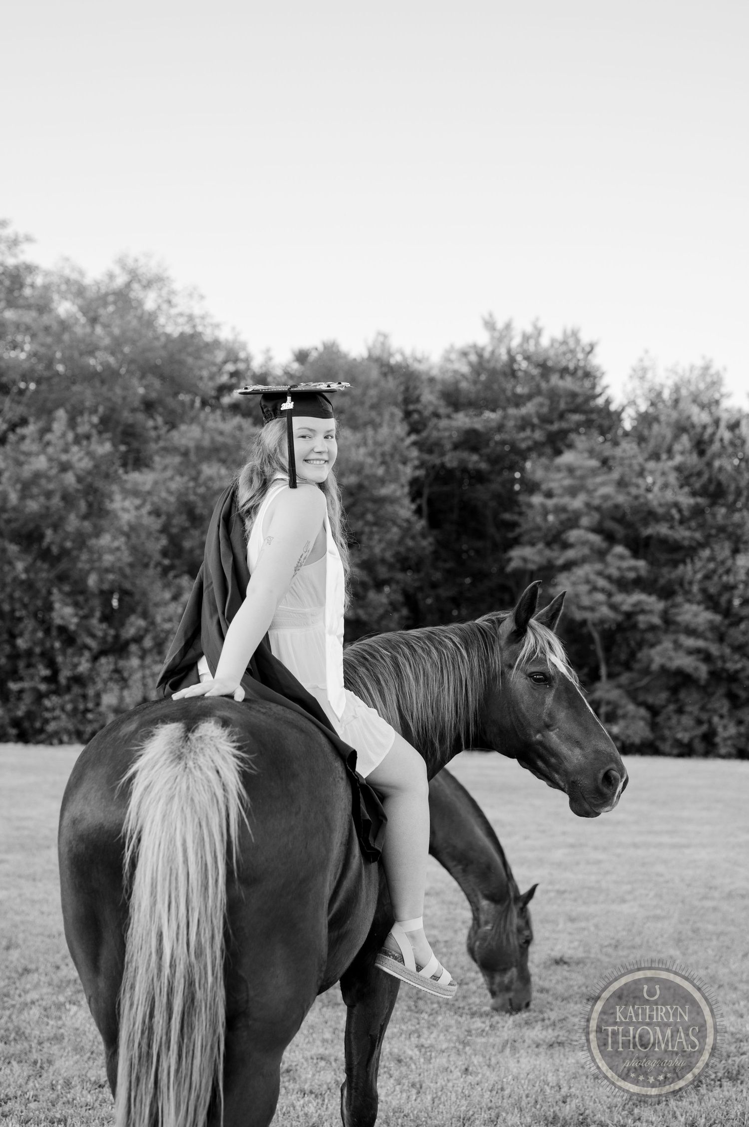 Black and white photo of a graduate in academic dress sitting sideways on a horse.