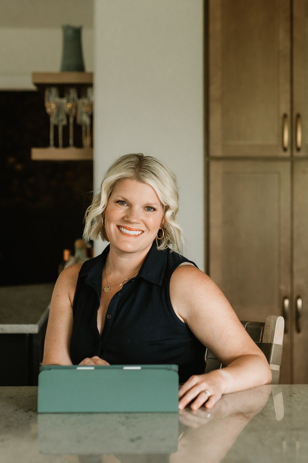Smiling blonde woman in black sleeveless top seated at a kitchen counter with a tablet.