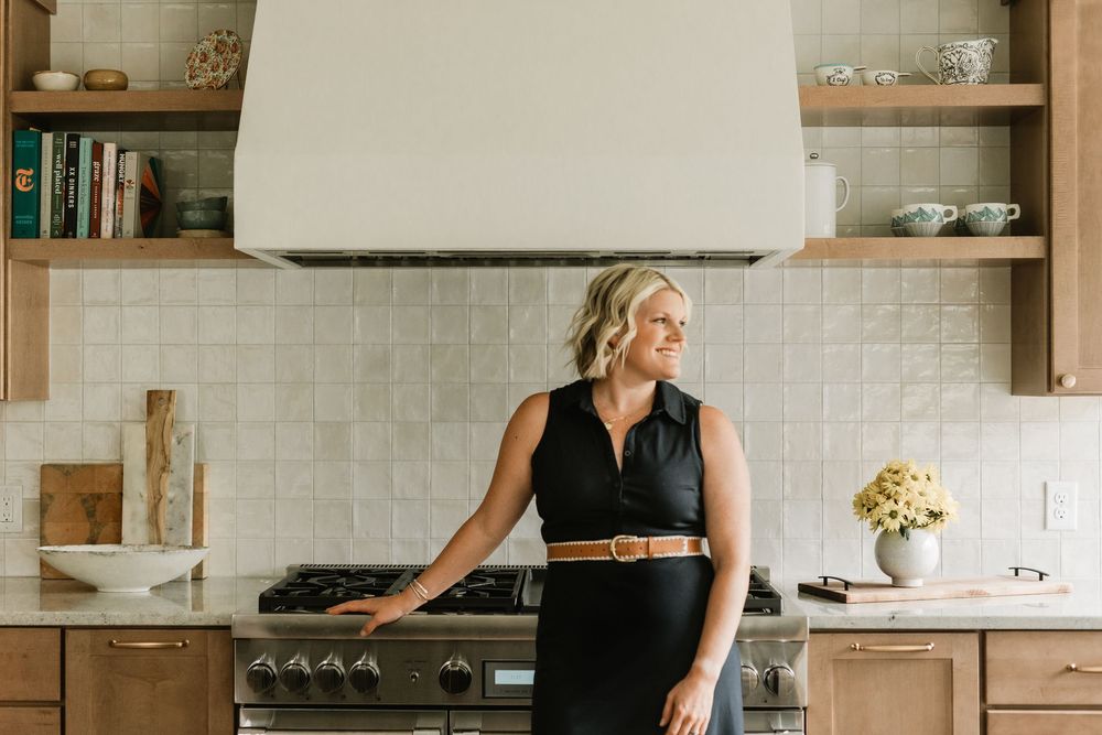 Woman in black dress standing in a modern kitchen with wood cabinets and stainless steel range.