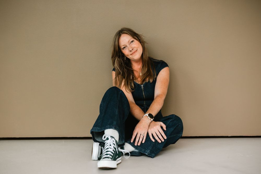 Smiling woman in black outfit and green Converse sneakers sitting casually on the floor.