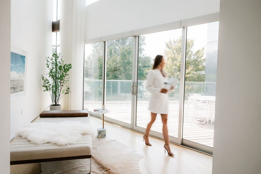 Woman in white dress standing by large floor-to-ceiling windows in a bright modern living space.