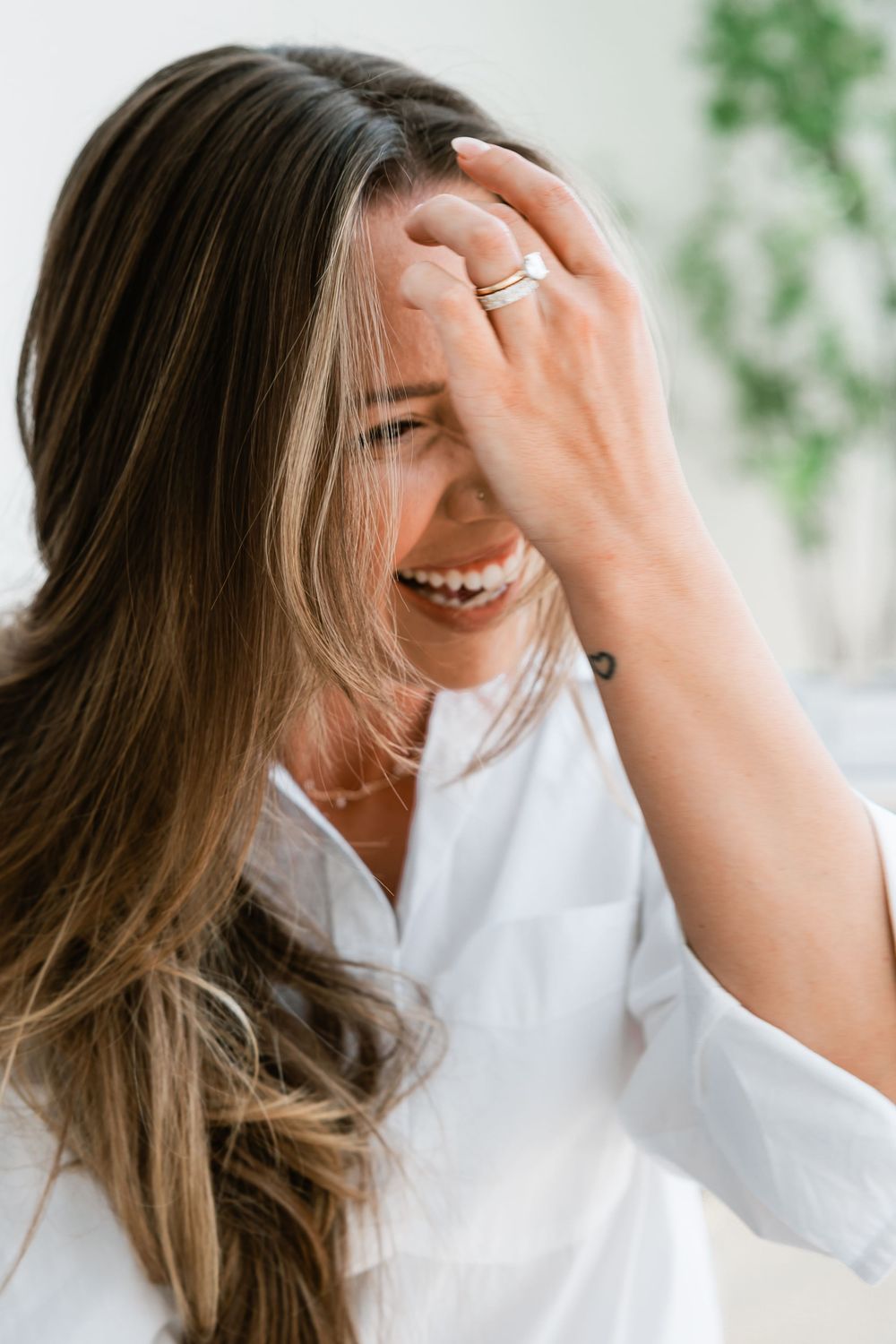 Laughing woman with long brown hair covering her face with her hand in a bright room.