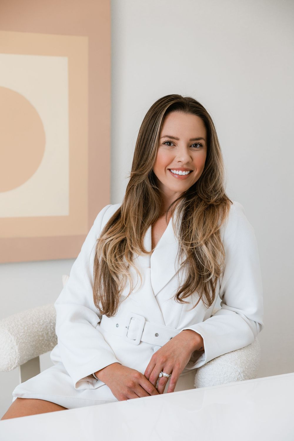Smiling woman with long brown hair wearing a white belted dress seated in a bright modern room.
