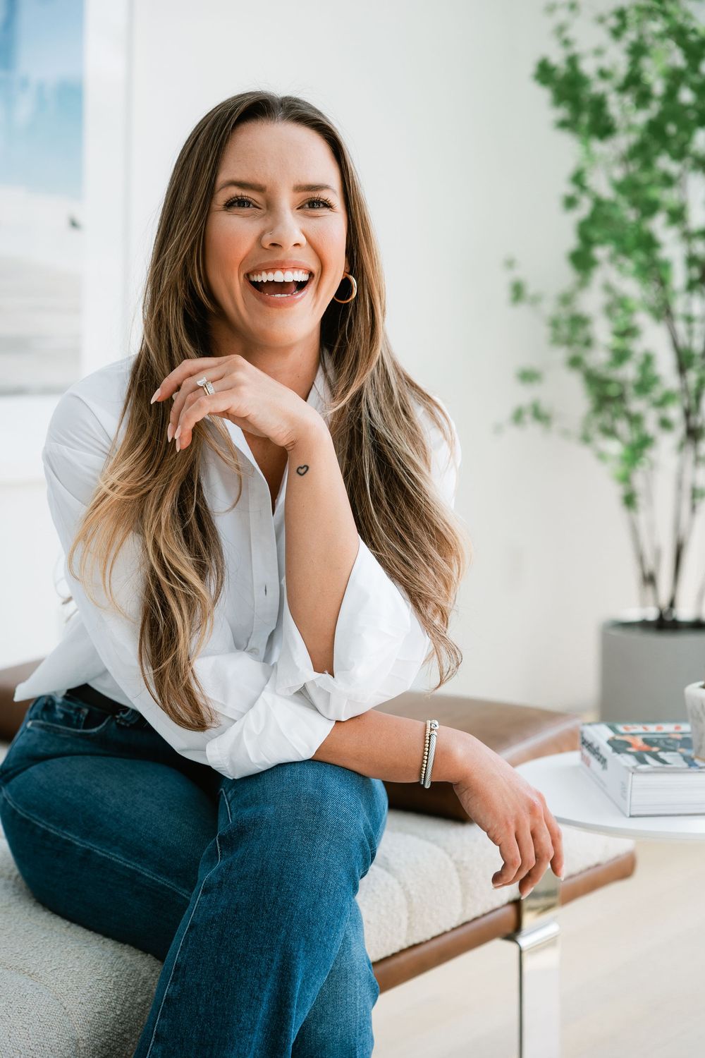 Cheerful woman with long brown hair in a white shirt and jeans laughing while seated indoors.