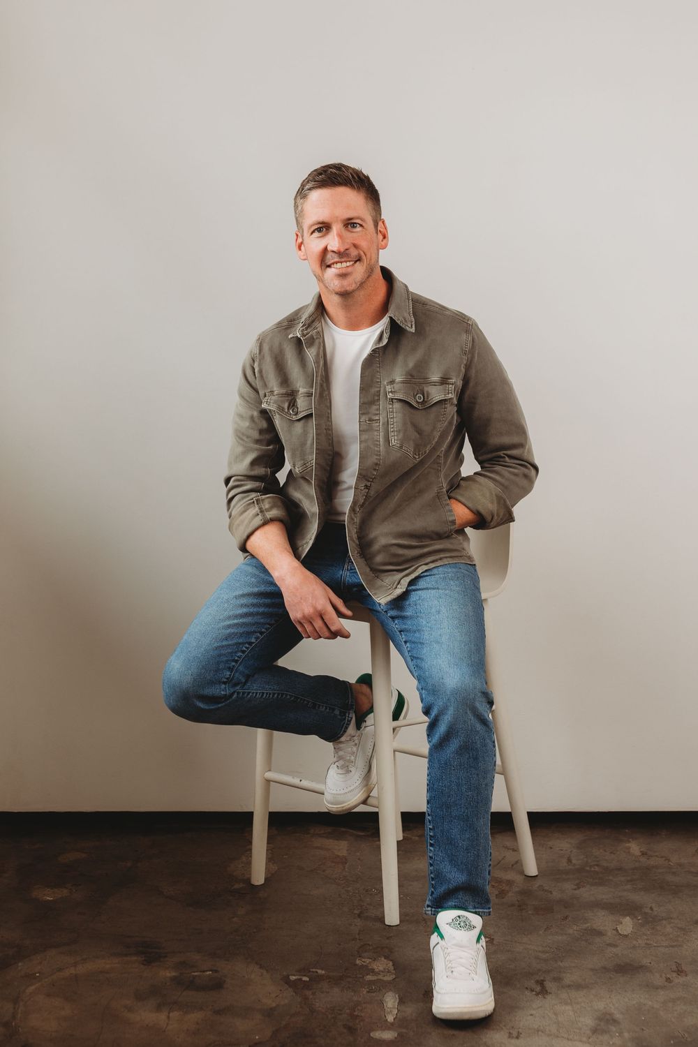 Smiling man in olive jacket and jeans sits relaxed on a white stool against a light gray wall.