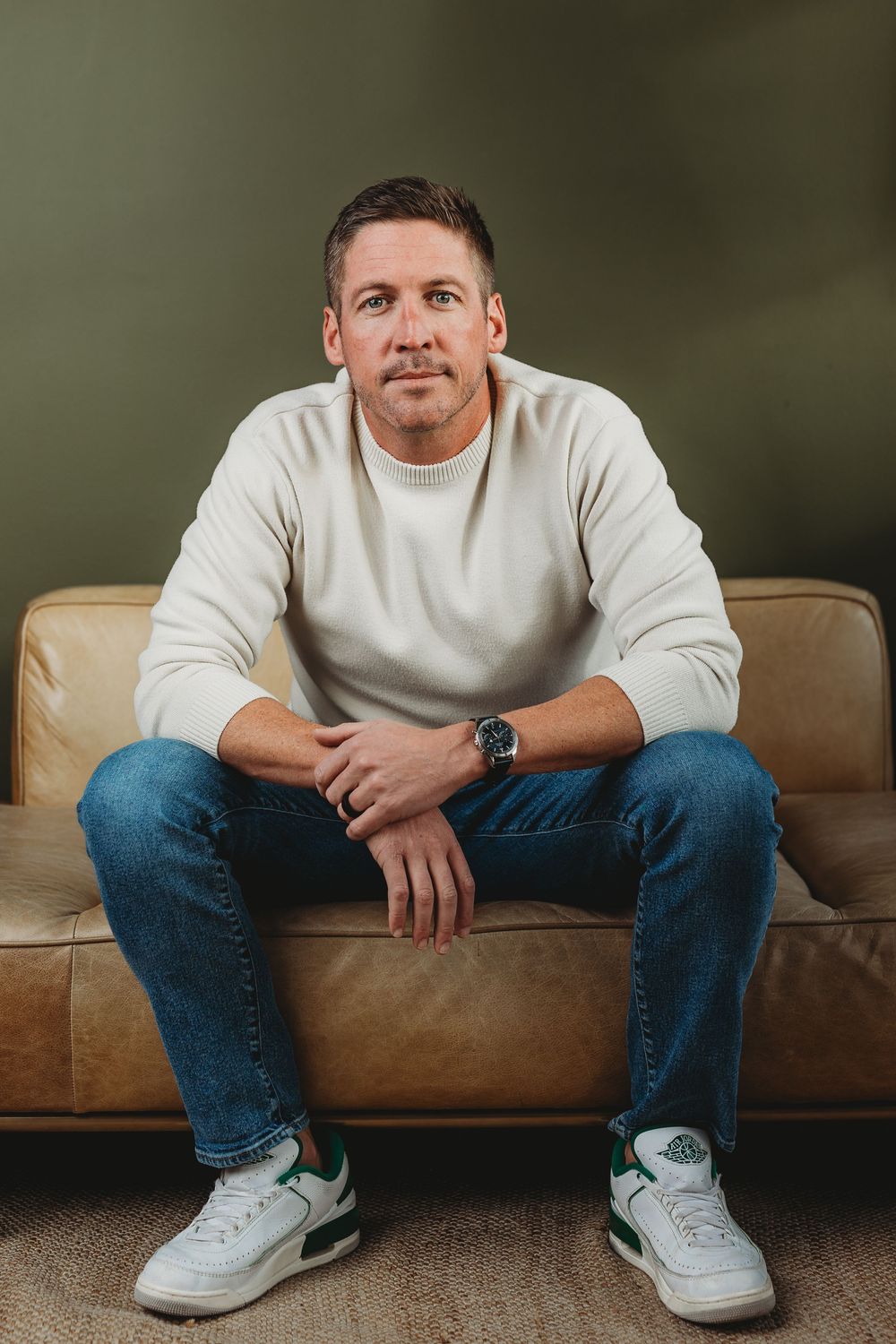 Confident man in white sweater and jeans sits on a leather sofa against an olive green wall.