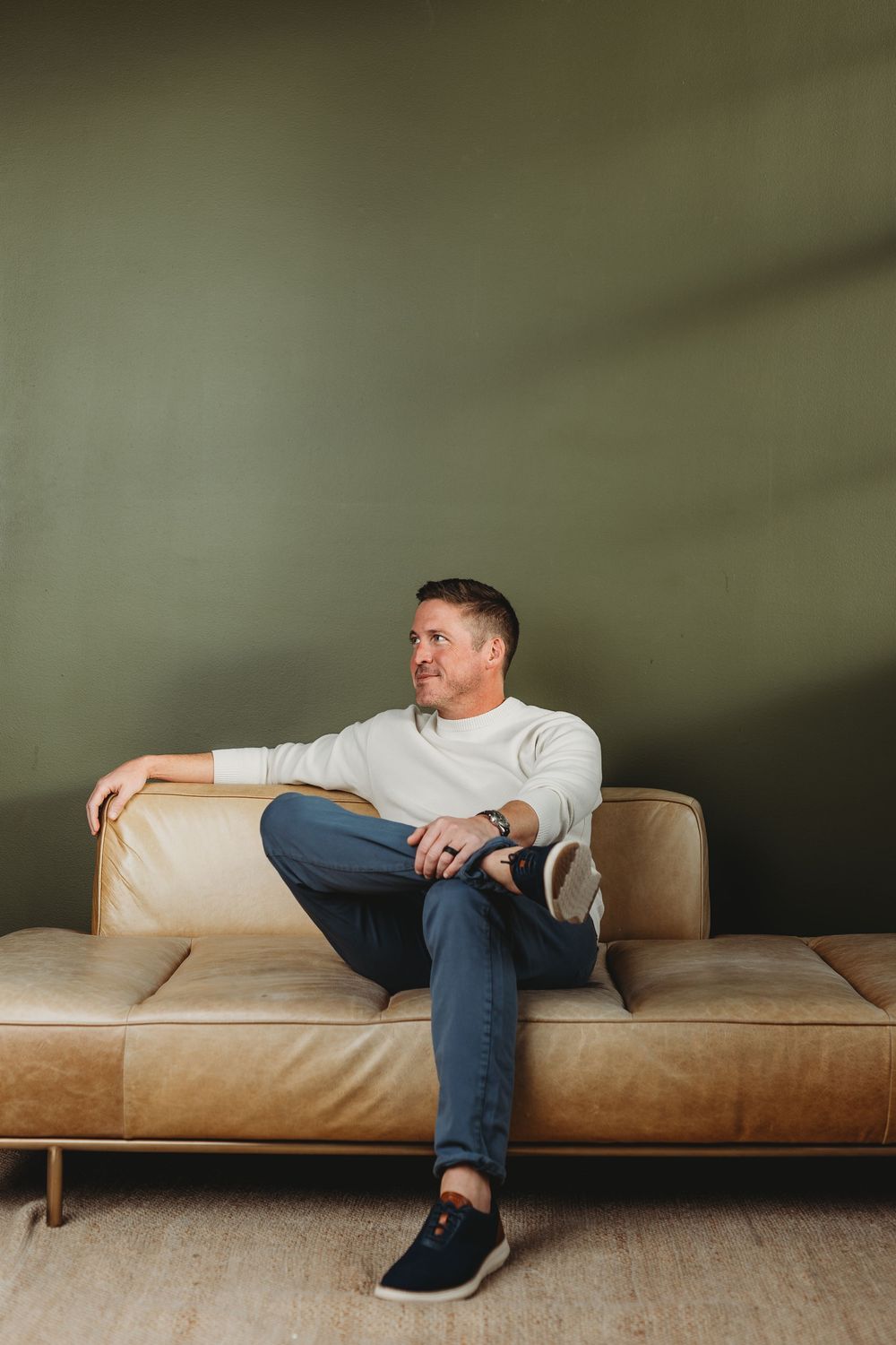 Man in white sweater and jeans sitting relaxed on a tan leather sofa against an olive green wall.