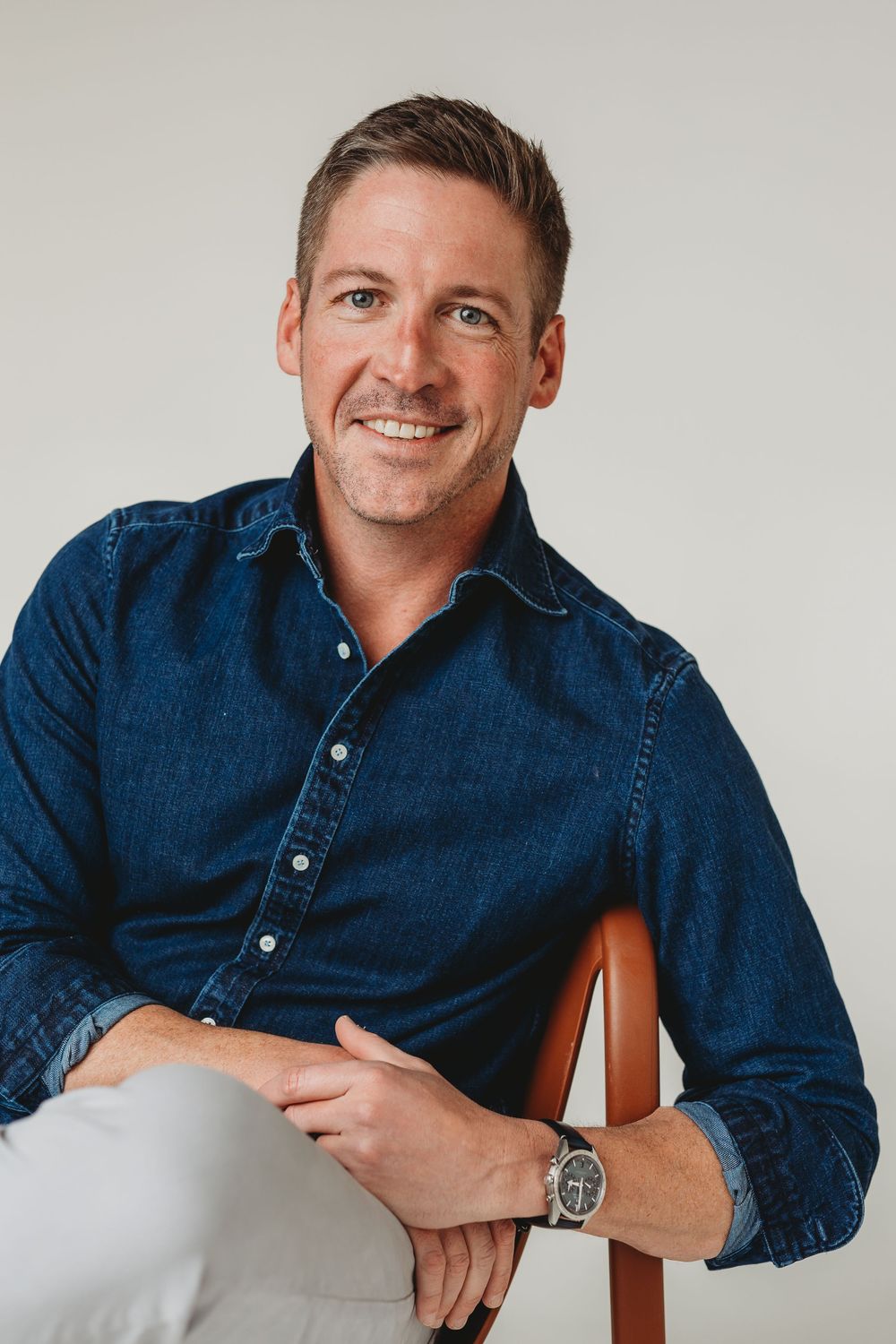 Smiling man in dark denim shirt seated on a chair against a light gray background.