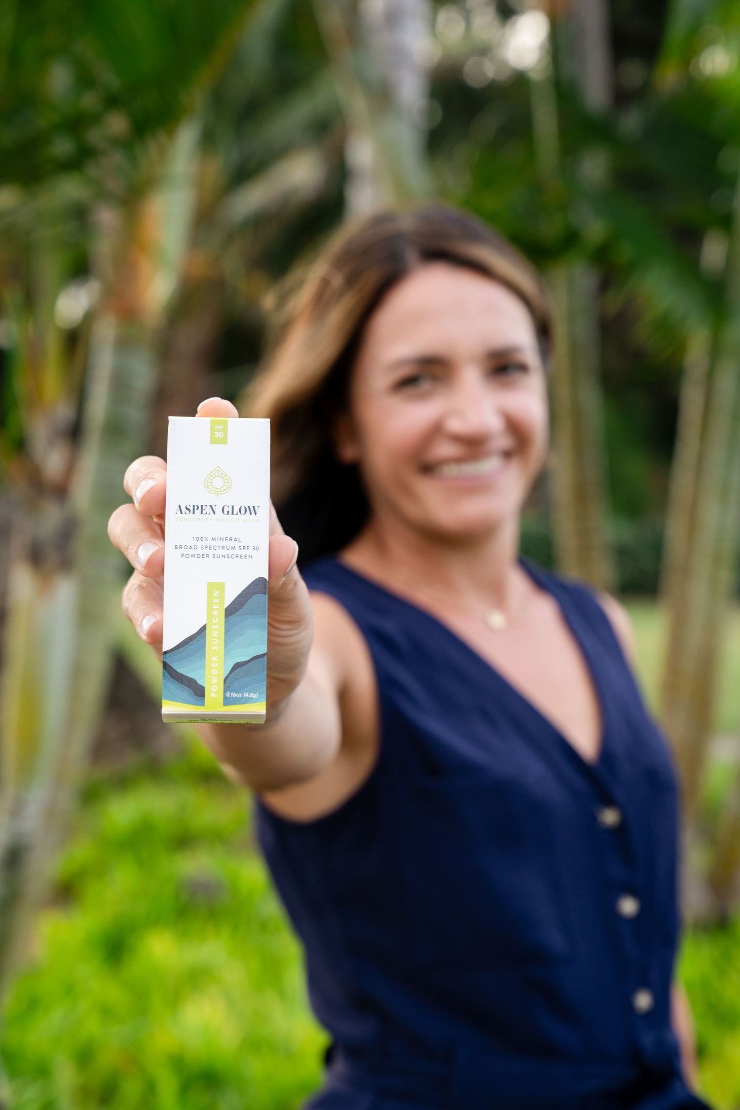 A person in a navy blue top holds up a white product box with green accents against a blurred natural background.