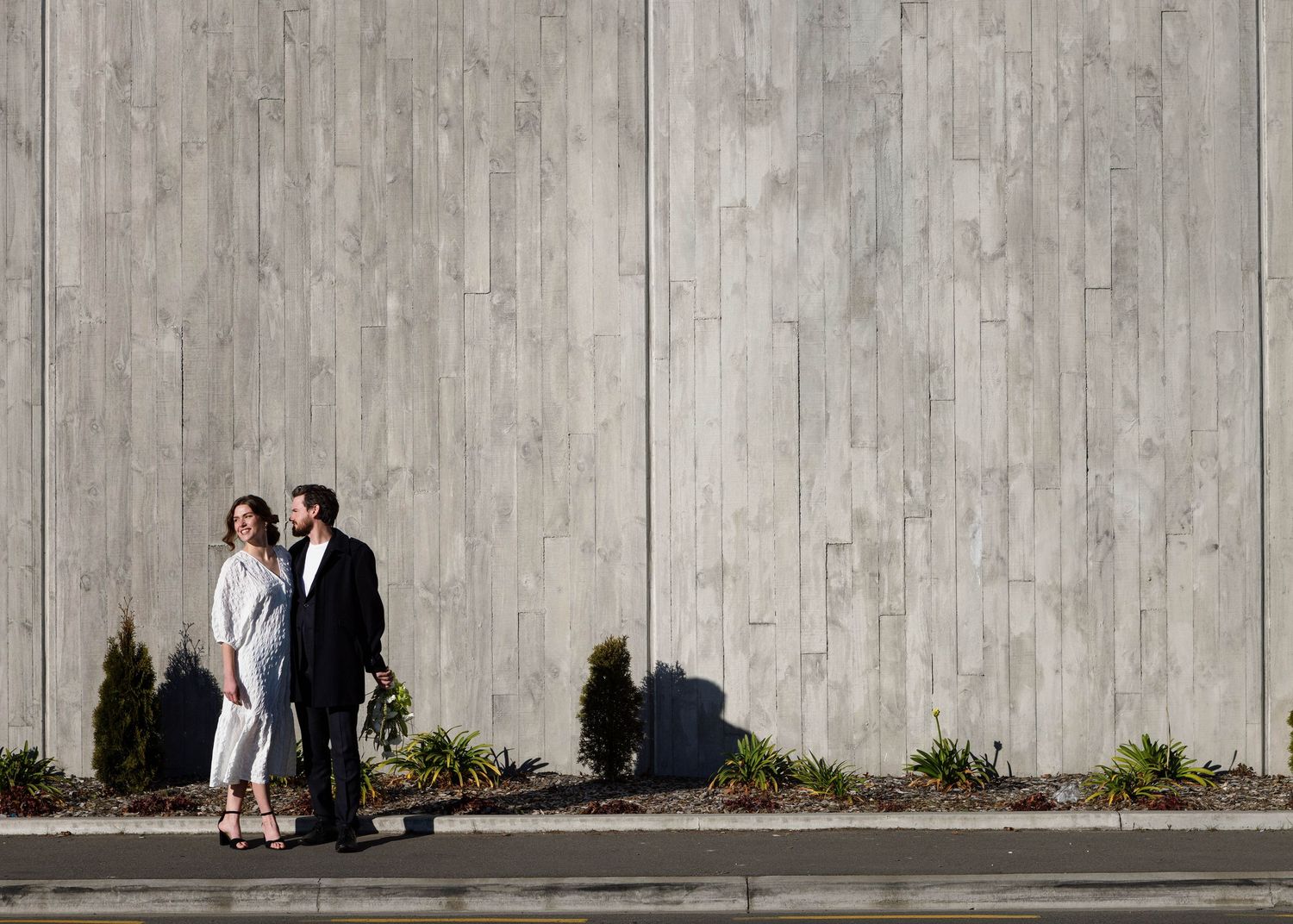 A couple stands against a concrete wall with evergreen shrubs on an urban sidewalk.