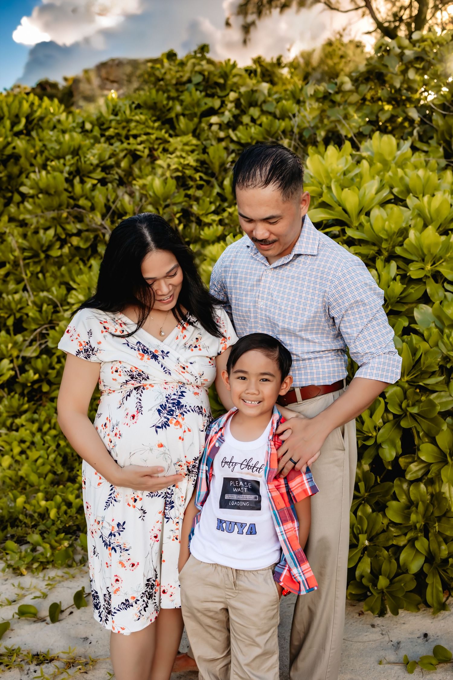 A pregnant woman in a floral dress stands with her family against lush green foliage on a sunny day.