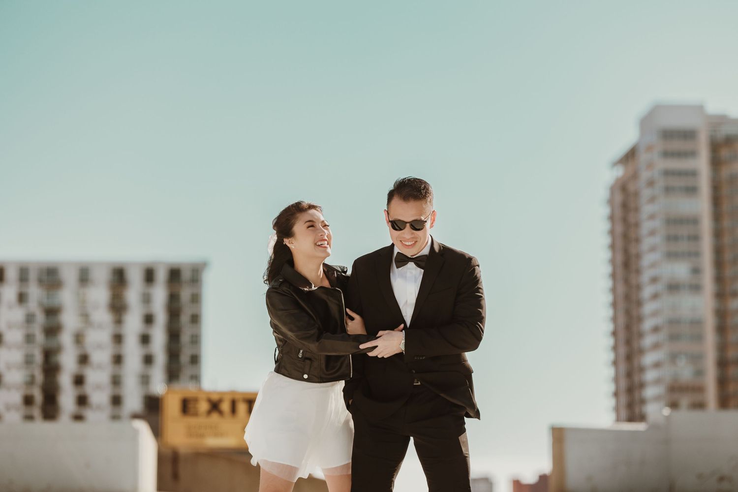 Stylish couple poses together on urban rooftop at sunset with cityscape and high-rise buildings in background.