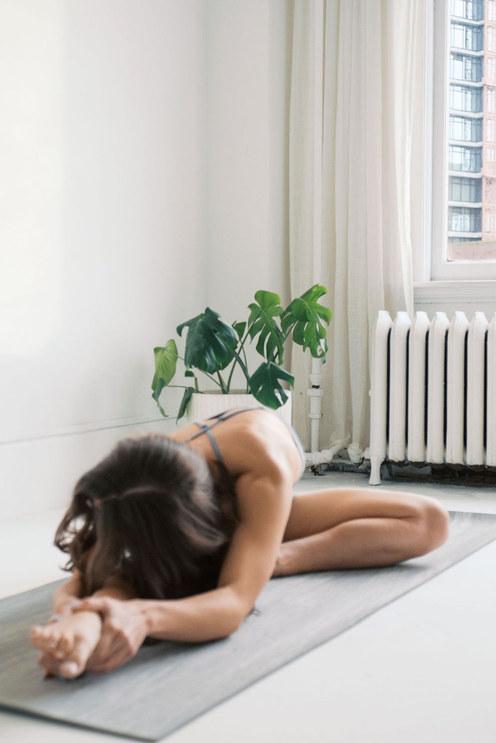 Yoga practitioner in resting pose on gray mat beside radiator and houseplant.
