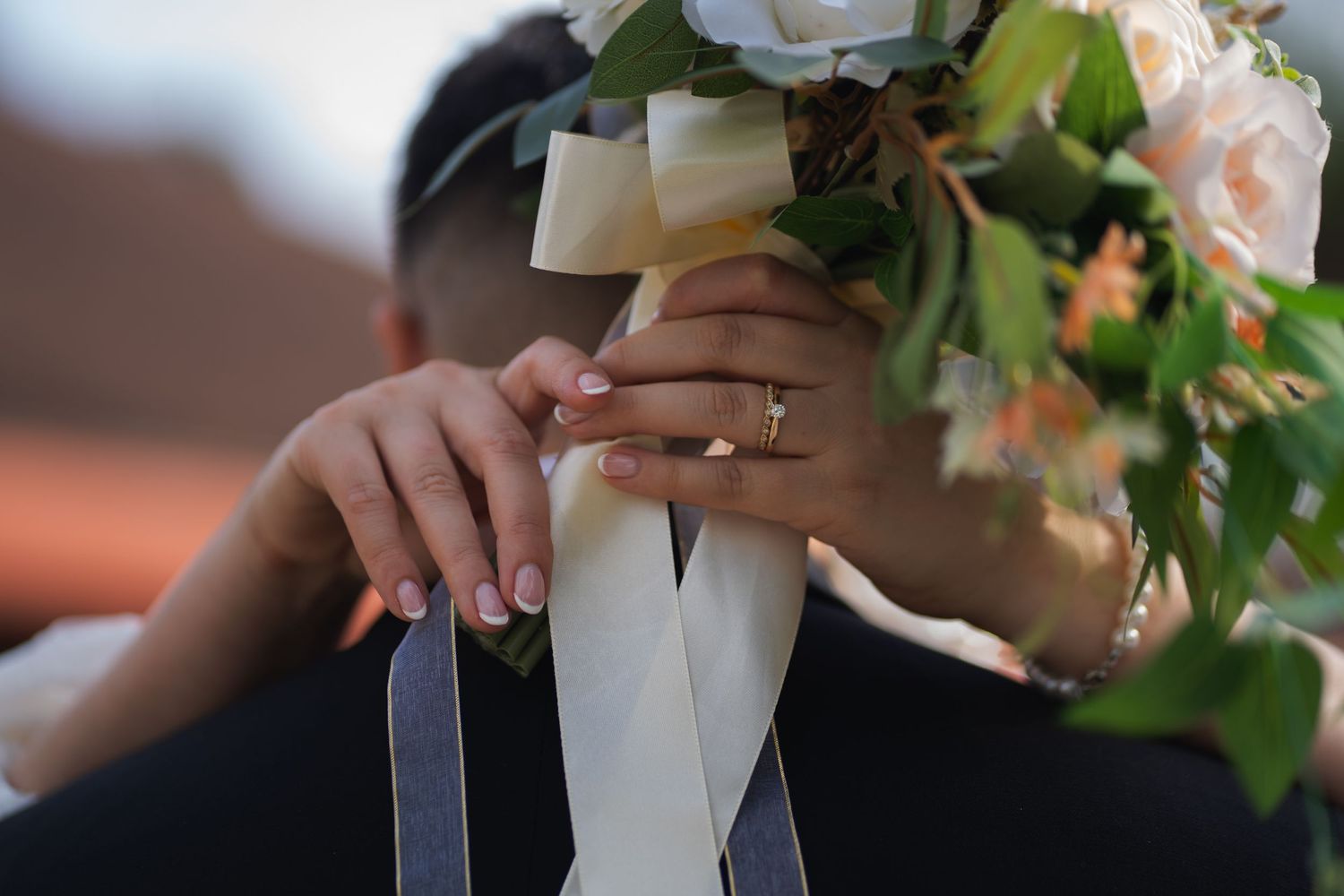Bride’s hands with engagement ring rest on groom’s shoulders, holding a floral bouquet. Elegant wedding photography, romantic bridal details