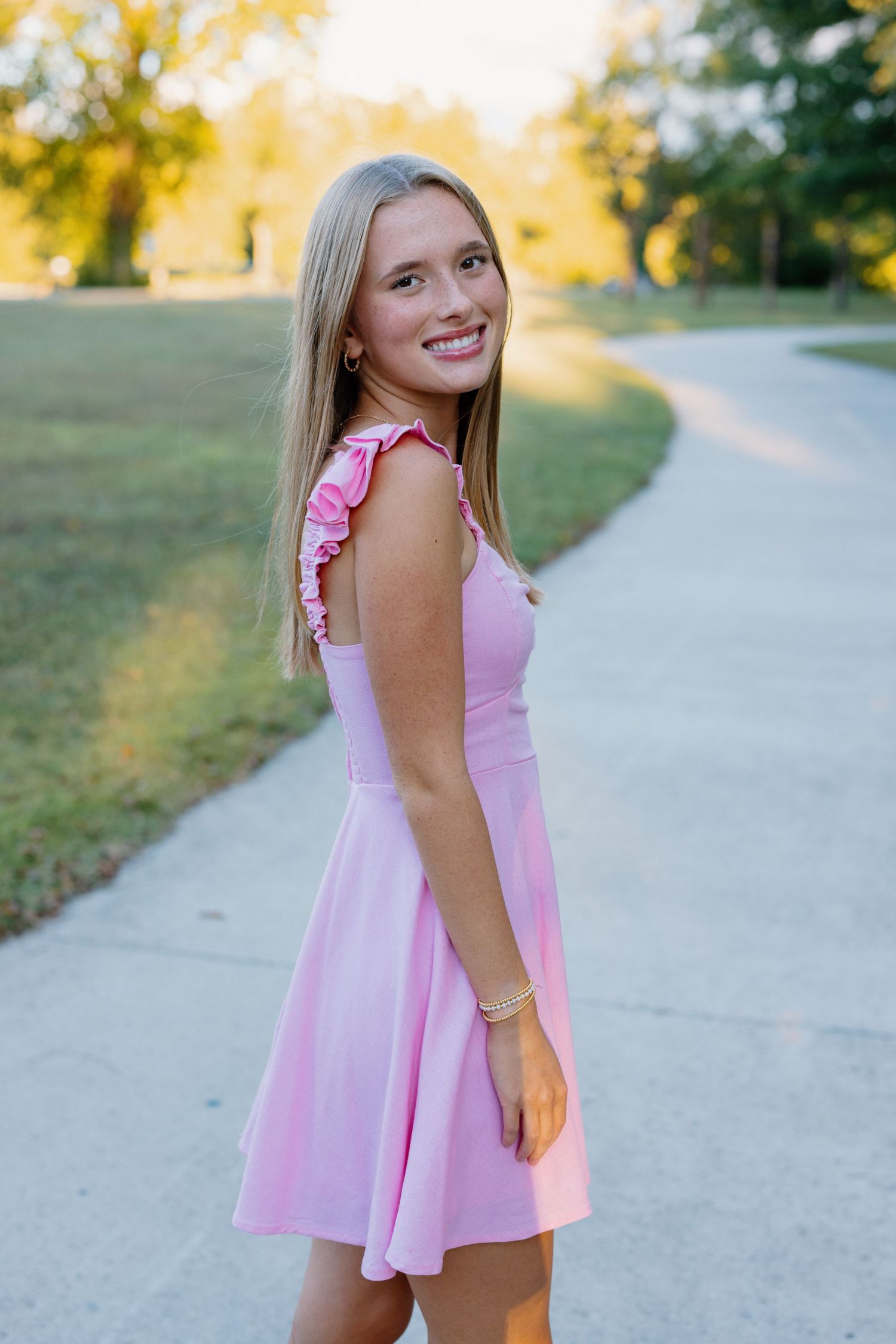 Young woman in pink dress smiling on outdoor path during golden hour.