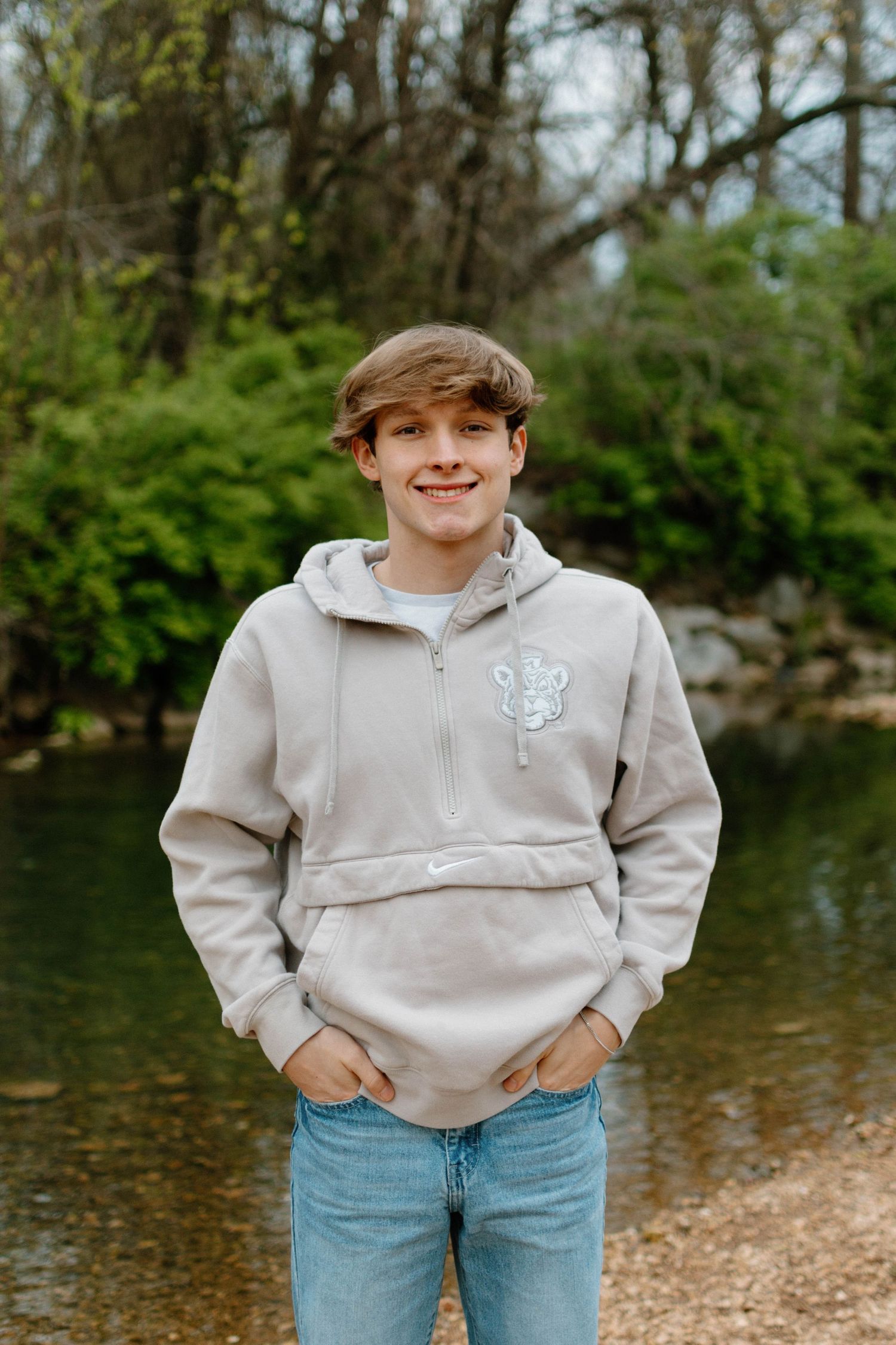 Young person in beige hoodie and jeans standing by water.