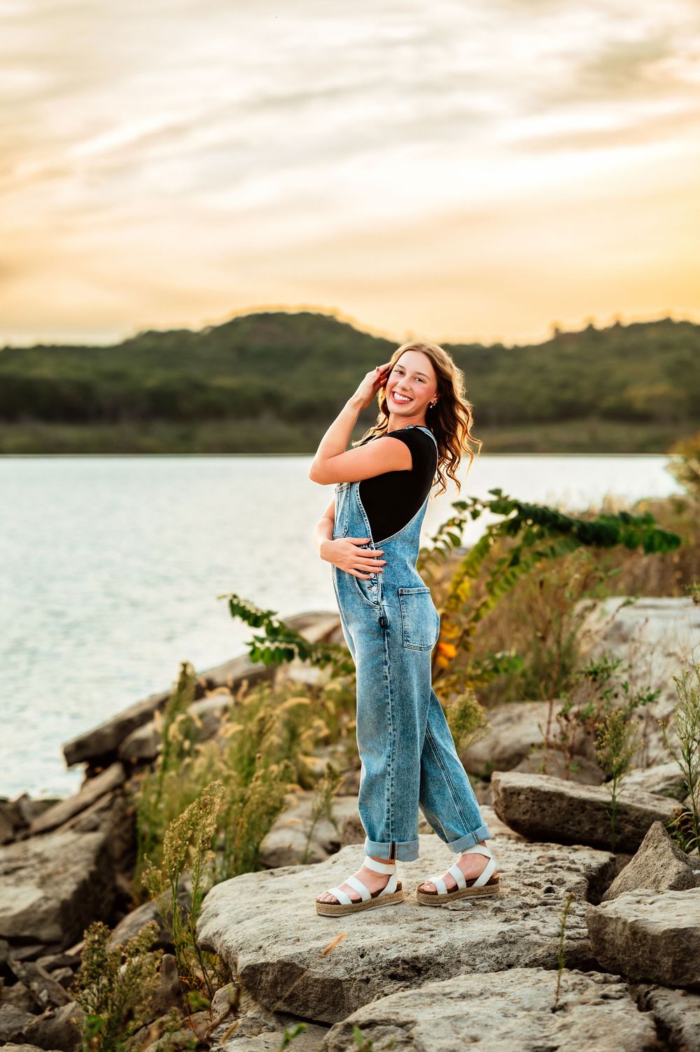 Person in casual denim and black top poses playfully on rocky shoreline at sunset by a lake.