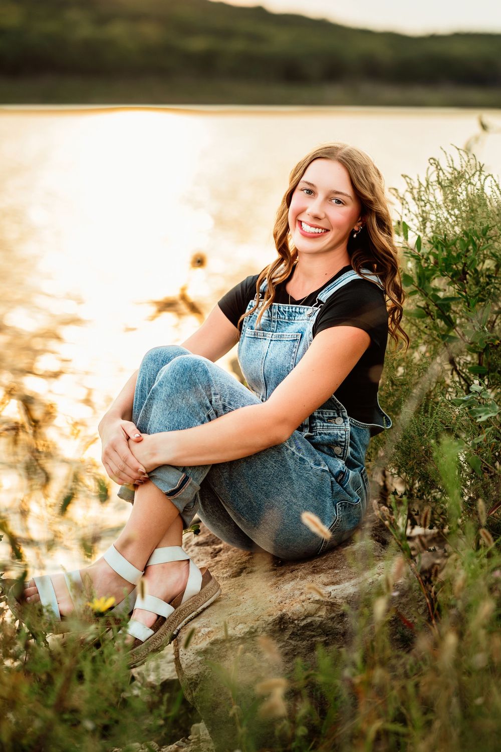 Outdoor portrait session in a field at golden hour featuring someone in denim overalls sitting on rocks.