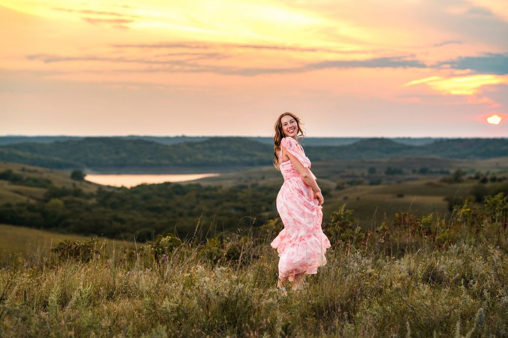Someone in a flowing pink dress twirls on a hilltop against a dramatic sunset landscape.