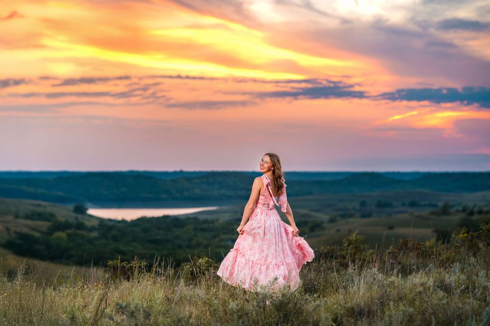 A flowing pink dress catches the breeze on a hilltop overlooking rolling countryside at sunset.