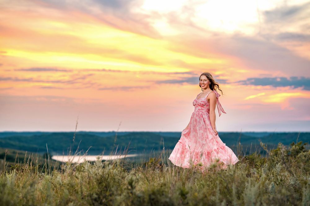 A dreamy sunset photo captures movement of a pink dress against a dramatic sky and rolling hills.