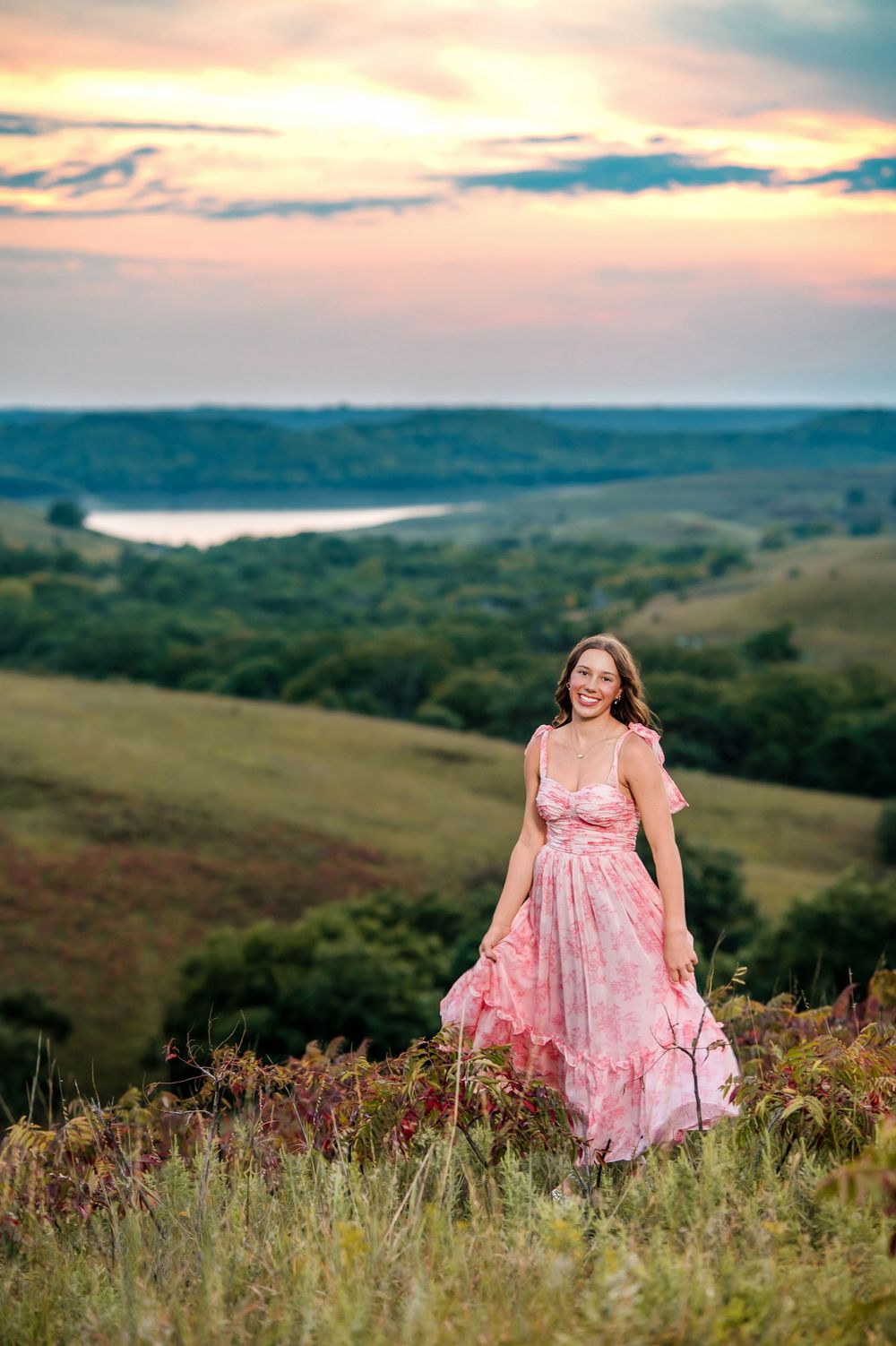 Scenic hilltop portrait in flowing pink dress with lake view at sunset.