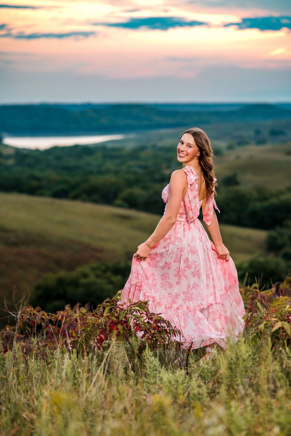 Scenic hilltop portrait in a pink floral maxi dress with rolling hills and lake in background at sunset.