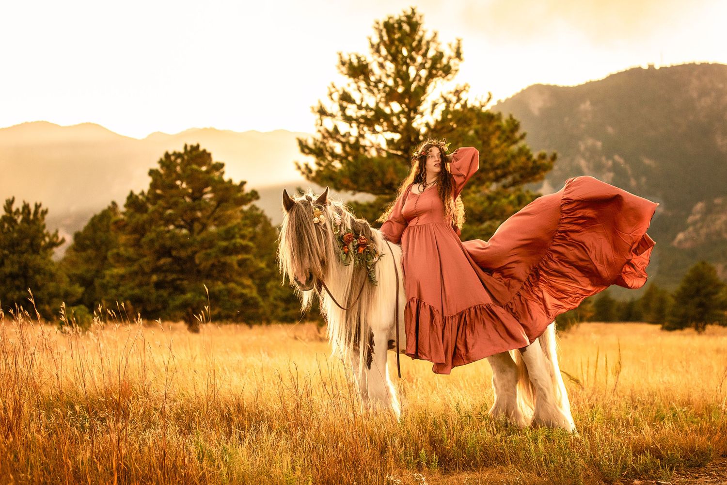 Woman in flowing red dress on white horse in golden mountain meadow at sunset.