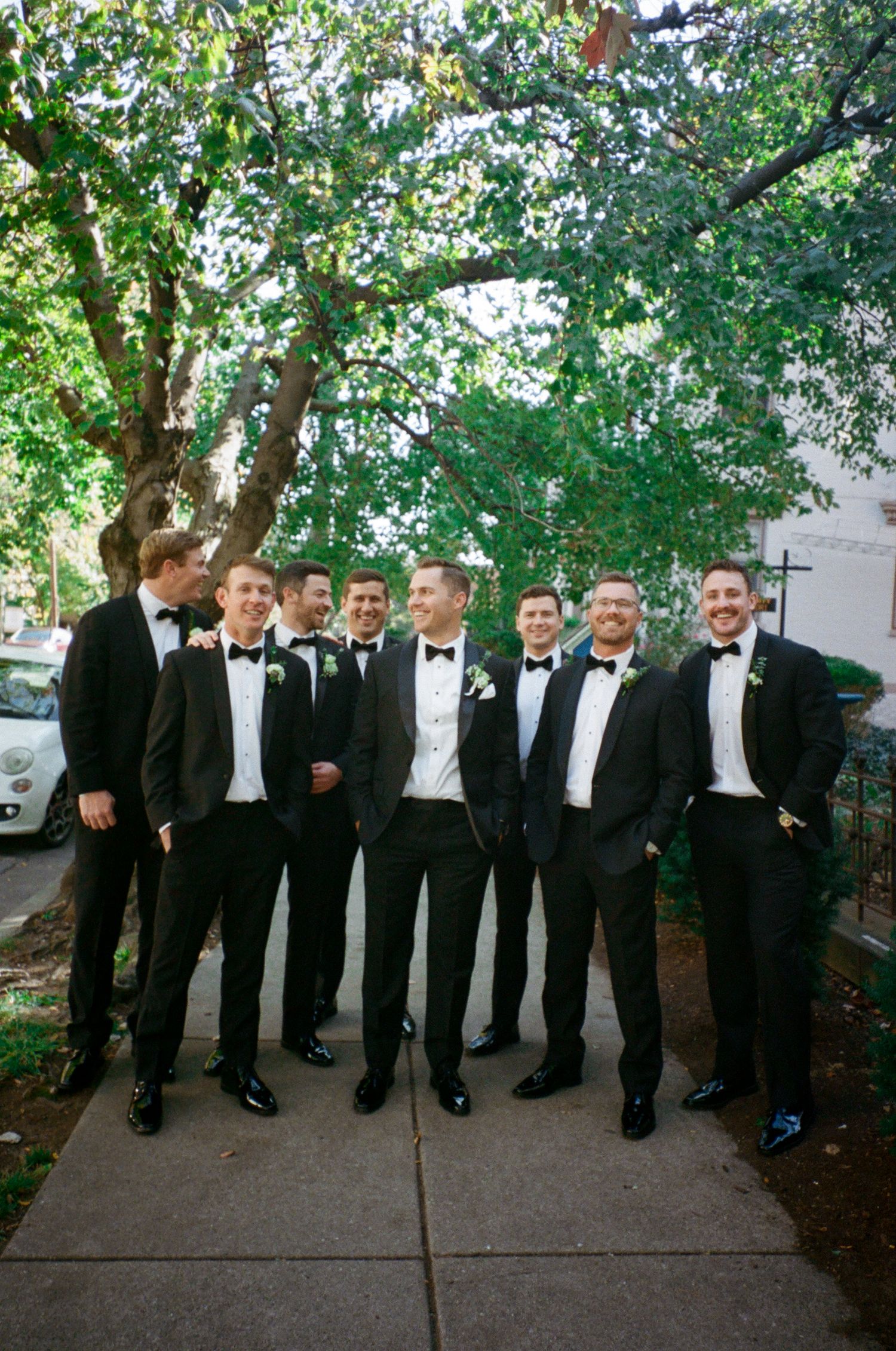 A group of groomsmen in black tuxedos pose together under trees.