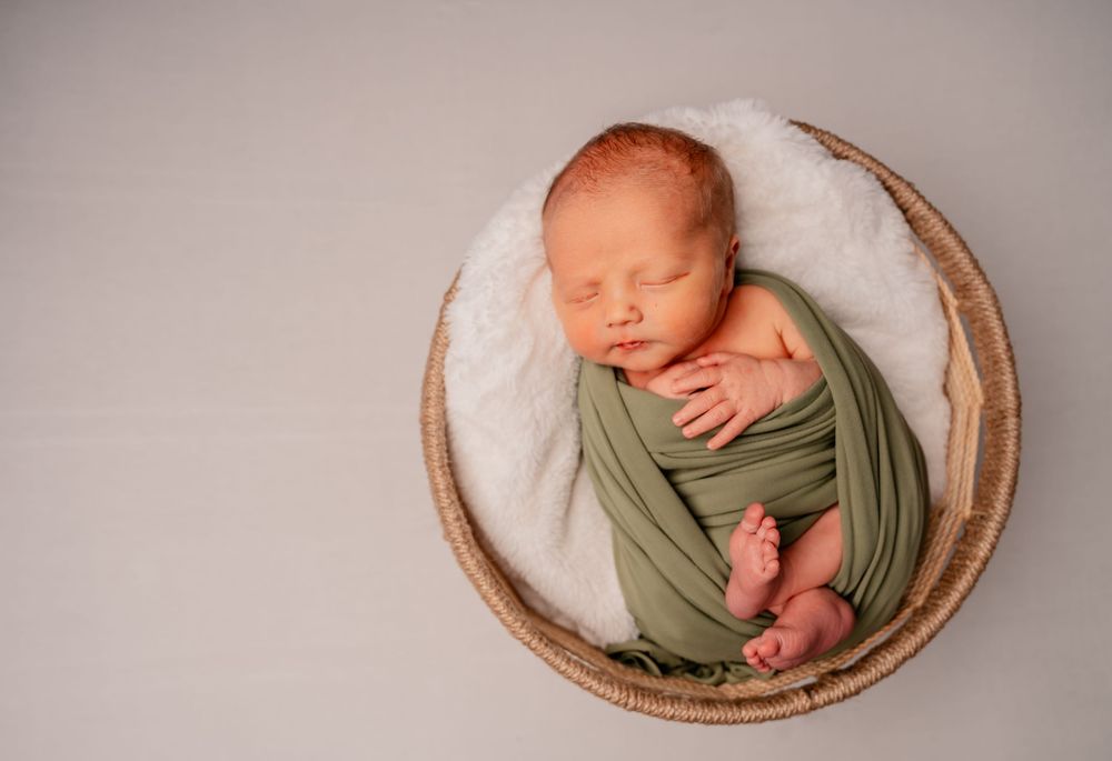 A sleeping newborn baby wrapped in sage green fabric nestles peacefully in a woven basket.