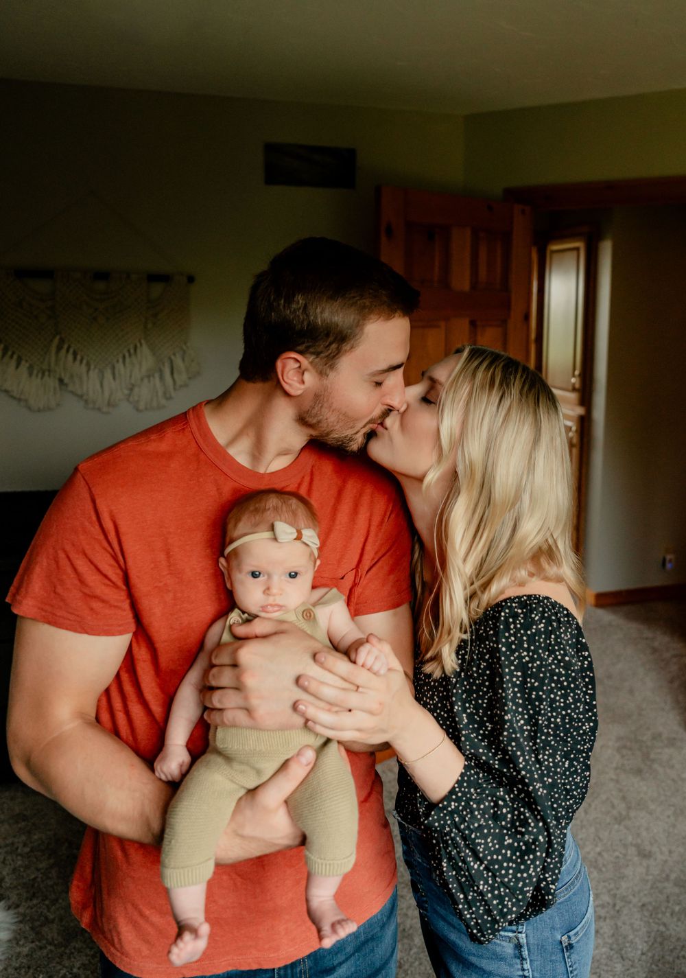 A joyful family moment with a baby in a tan onesie being held between loving parents.
