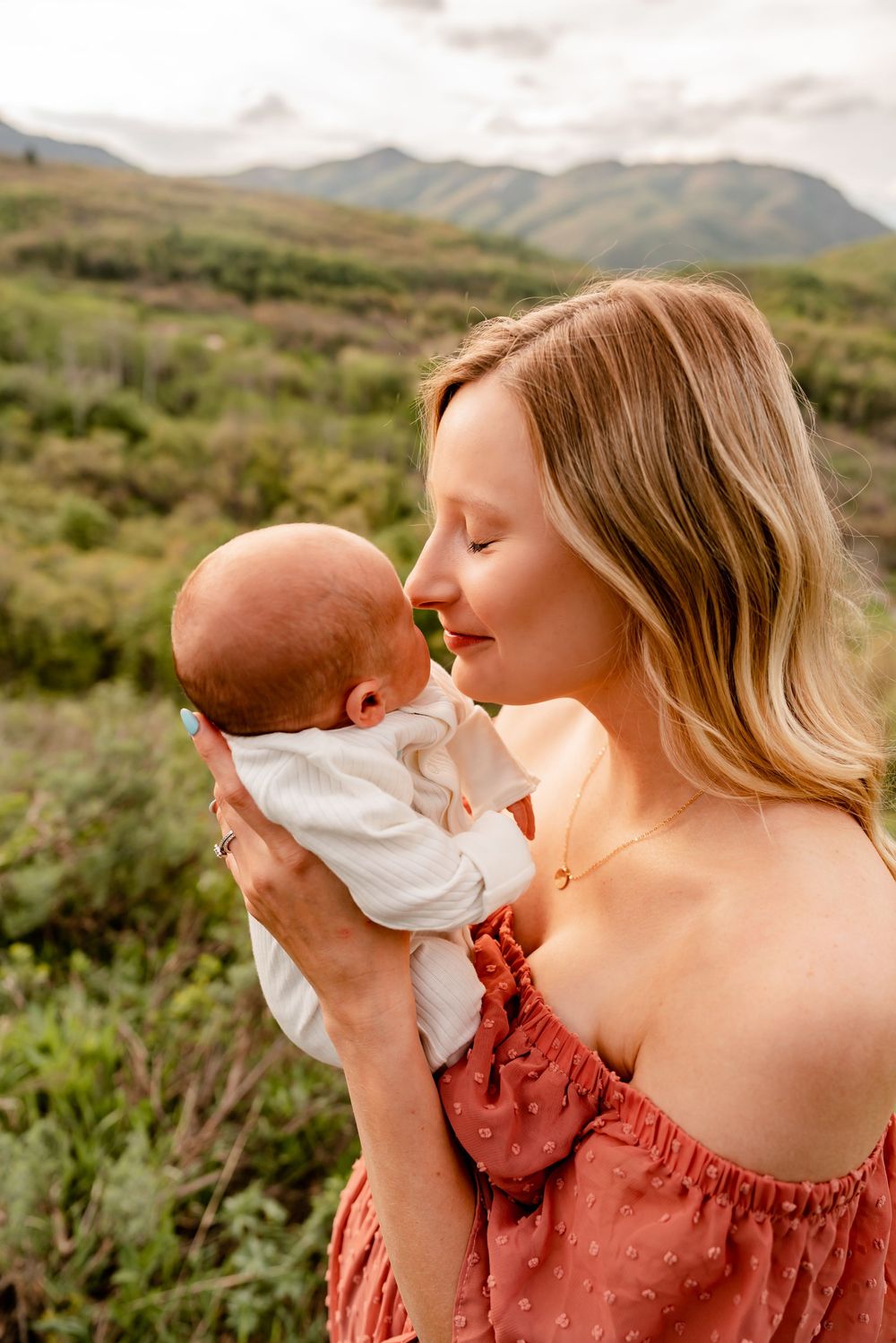 Tender moment shared between mother and infant against backdrop of rolling mountain hills at sunset.