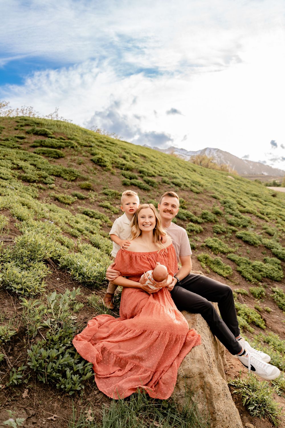 A group relaxes together on a grassy mountain hillside beneath cloudy skies wearing casual attire with warm earth tones.