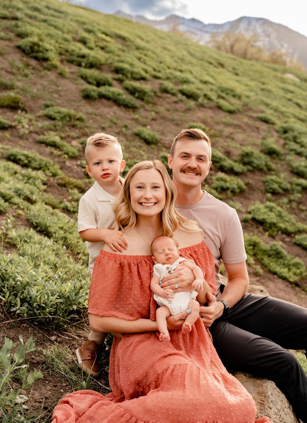 A family in coordinated outfits poses together on a mountain trail with green slopes and cloudy skies in the background.