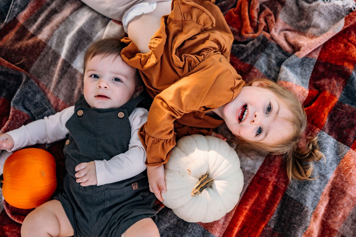 Children lying on blanket with orange and white pumpkins in autumn setting.
