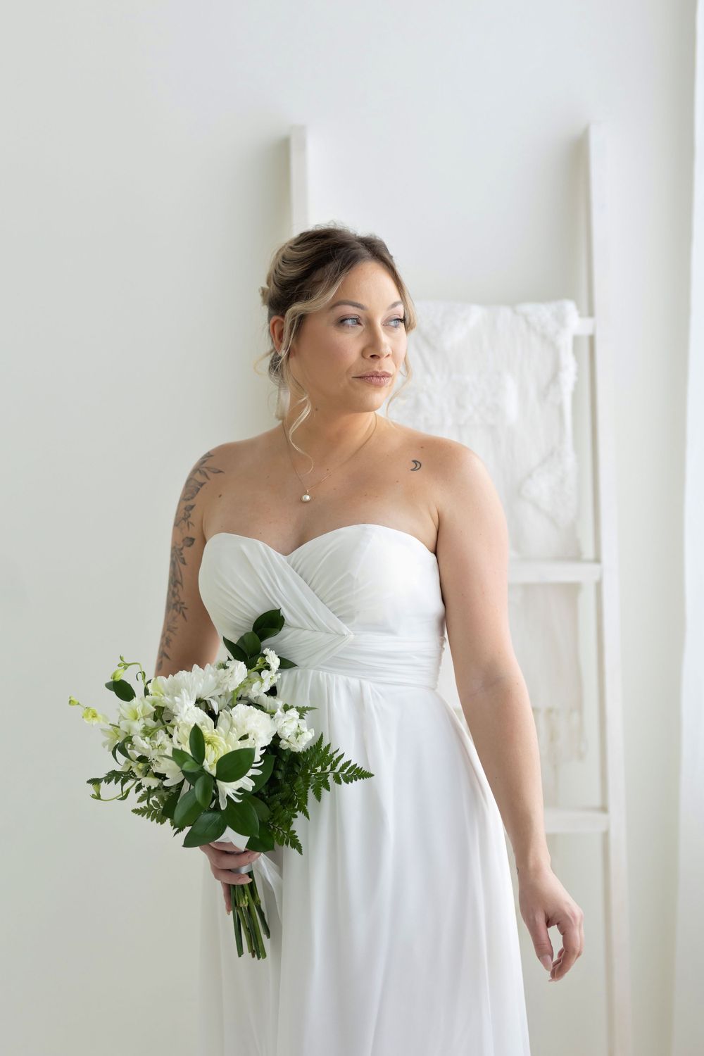 Tattooed bride in strapless white dress holding white flower bouquet standing near decorative ladder.
