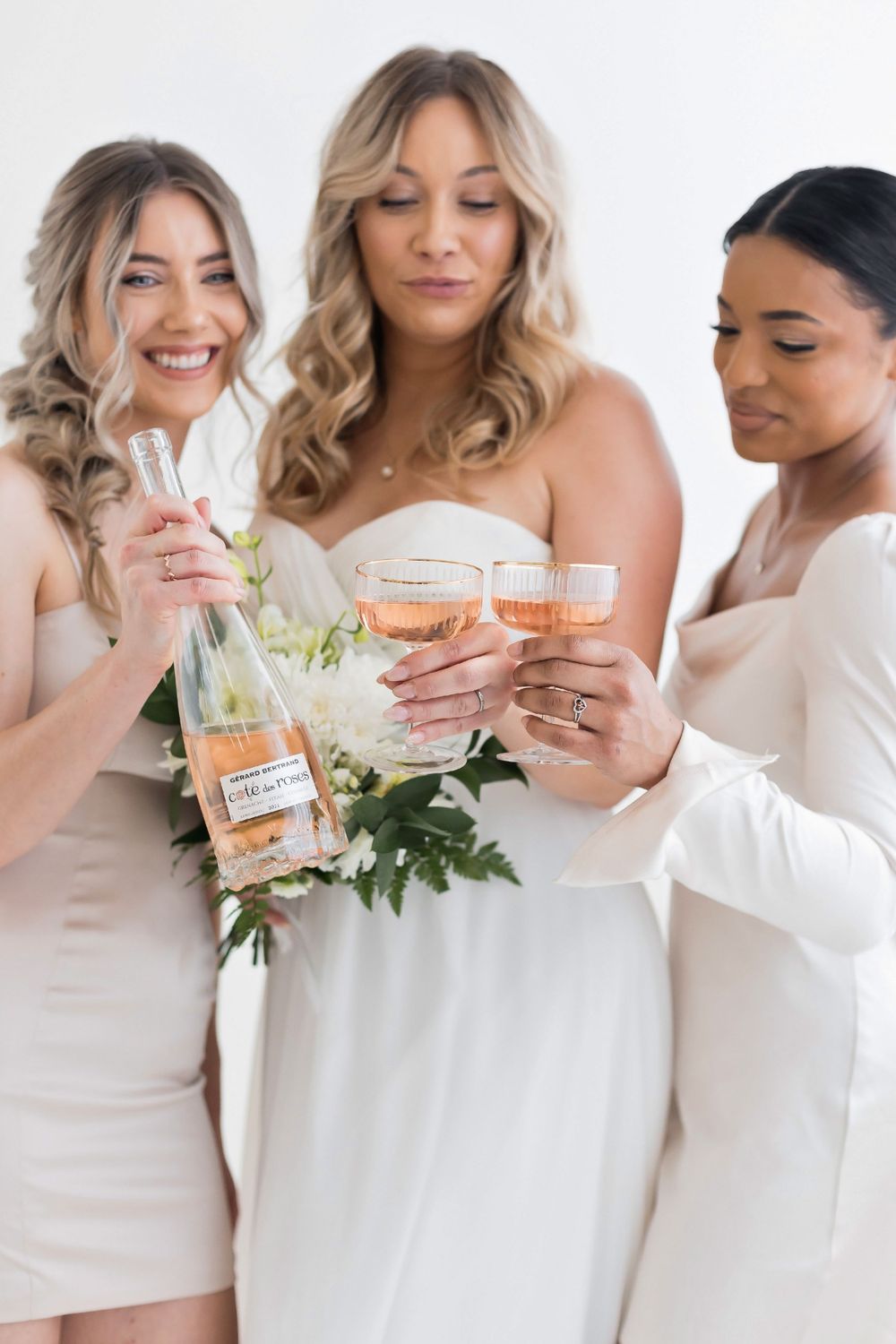 Bride and two bridesmaids in white dresses toasting with rose champagne glasses and bottle.