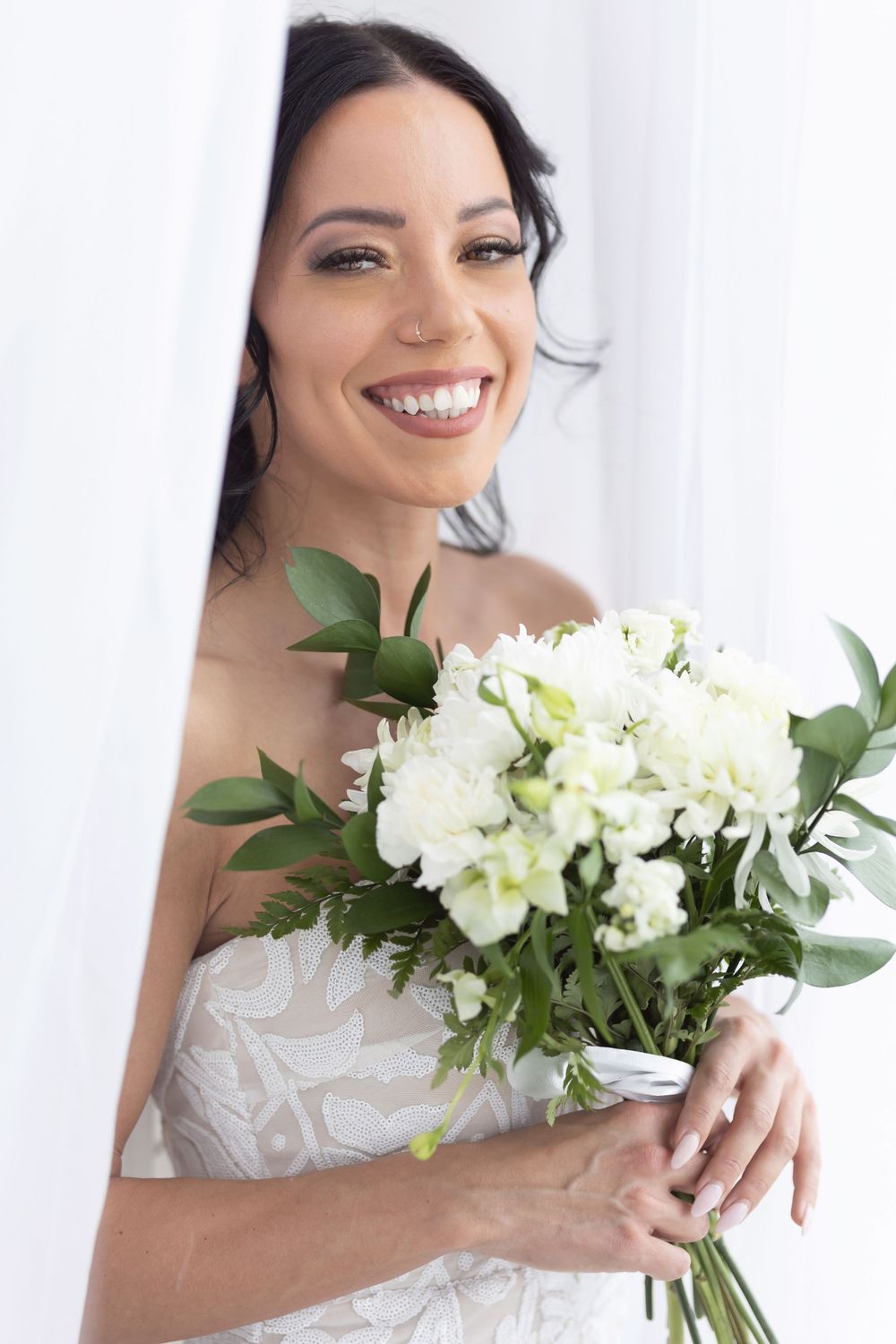 Smiling dark-haired bride in white lace dress holding white flower bouquet near curtains.