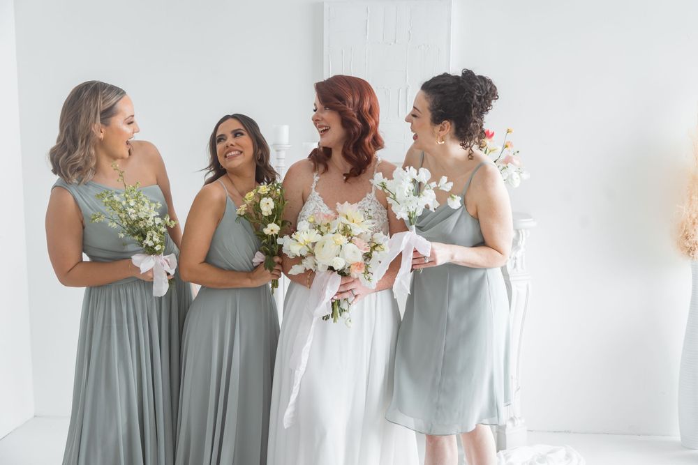Bride with bridesmaids in sage green dresses holding white floral bouquets laughing together indoors.