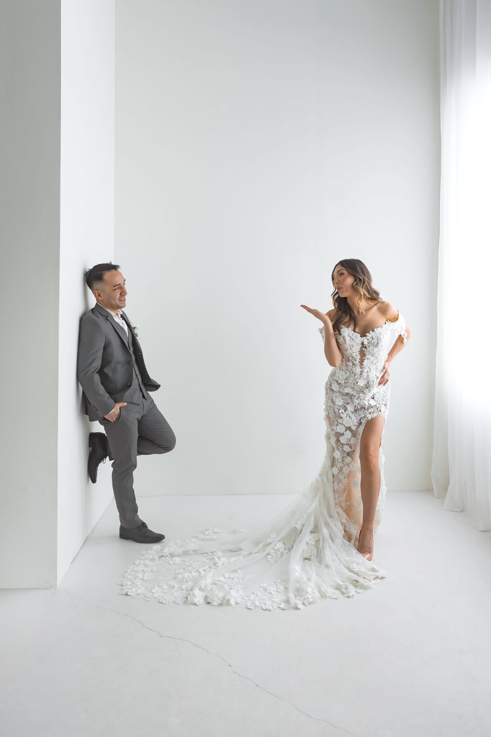 Bride in floral lace wedding dress and groom in gray suit posing playfully against white studio wall.