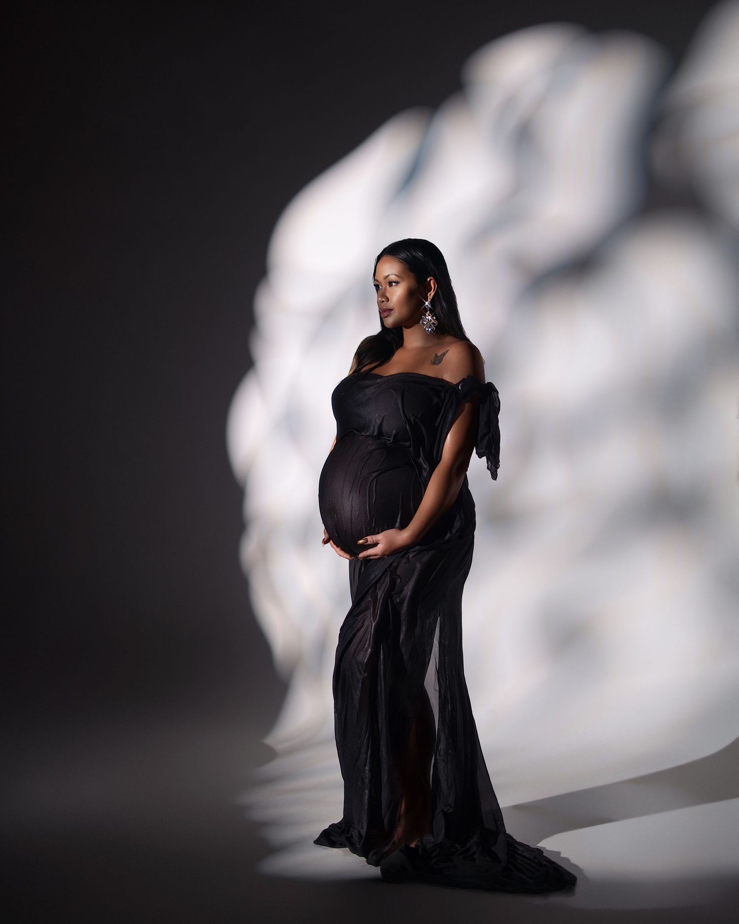 Elegant pregnant woman in black off-shoulder gown posing for maternity photo shoot against dramatic light backdrop.