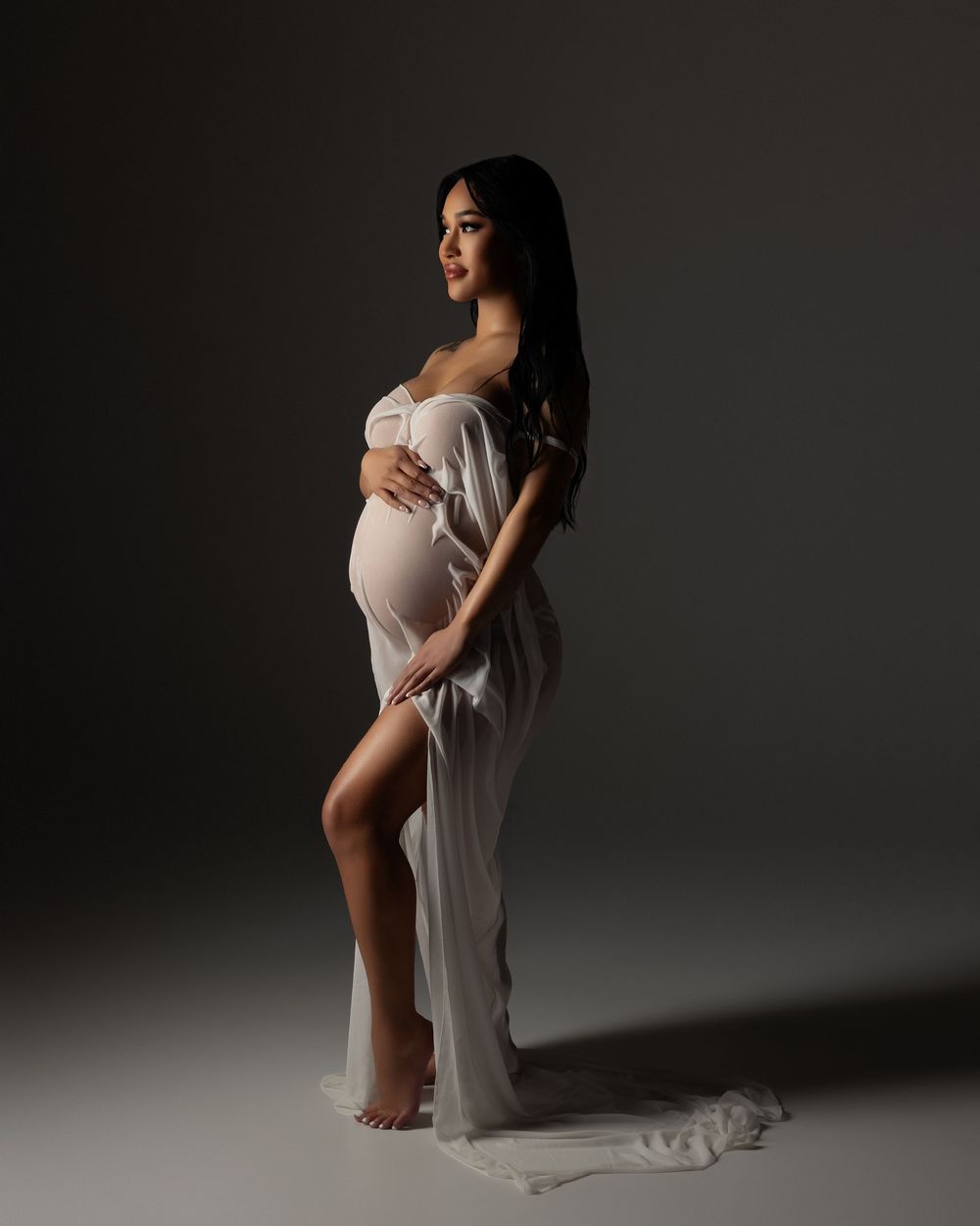 Artistic maternity portrait showing an elegant silhouette in flowing white fabric against a dark studio backdrop.